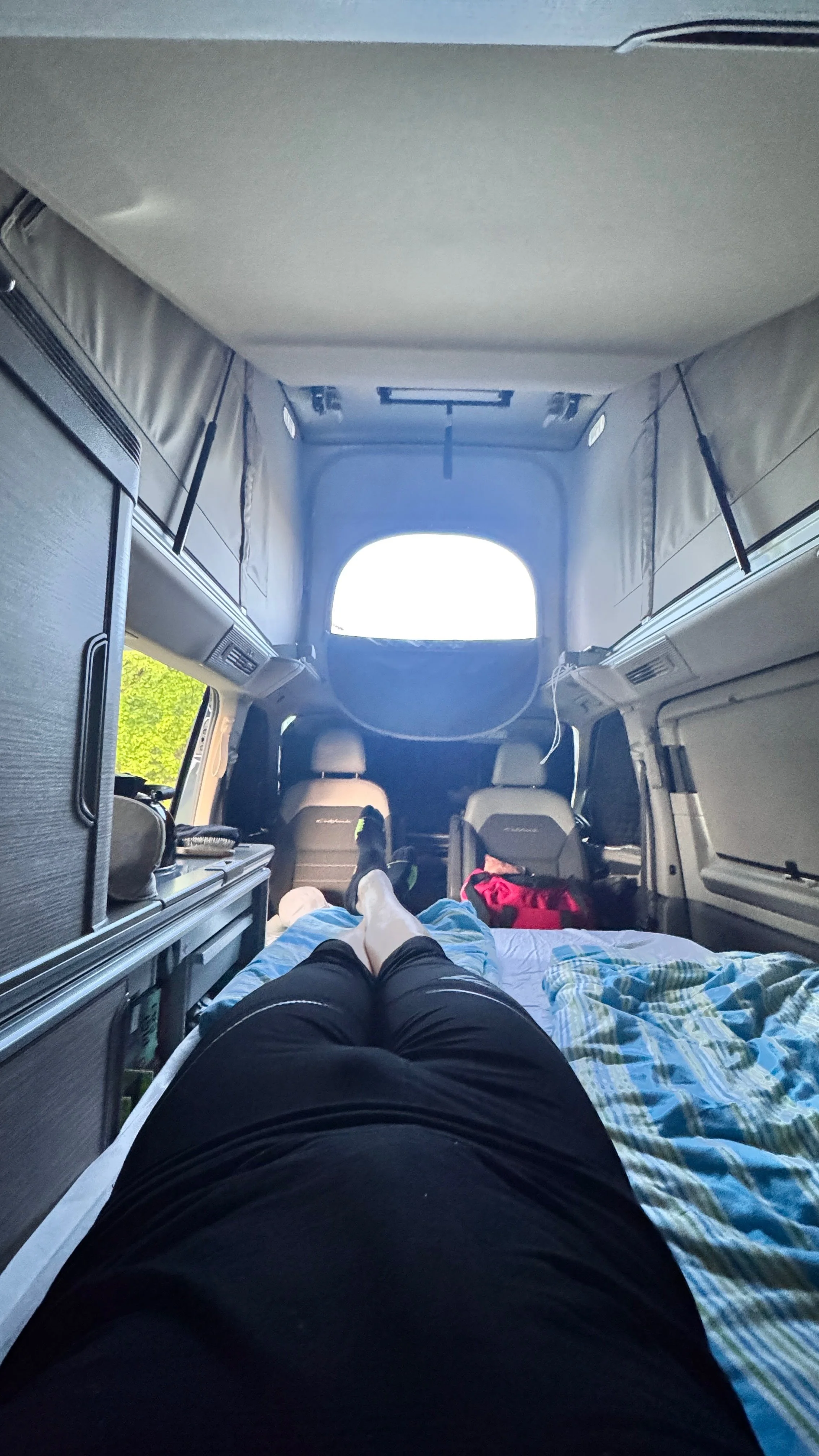 Inside a camper van, a person is lying on a bed with their legs stretched out, wearing black shorts and white socks. The front seats and windshield of the camper are visible, with a window open at the back showing bright daylight outside.