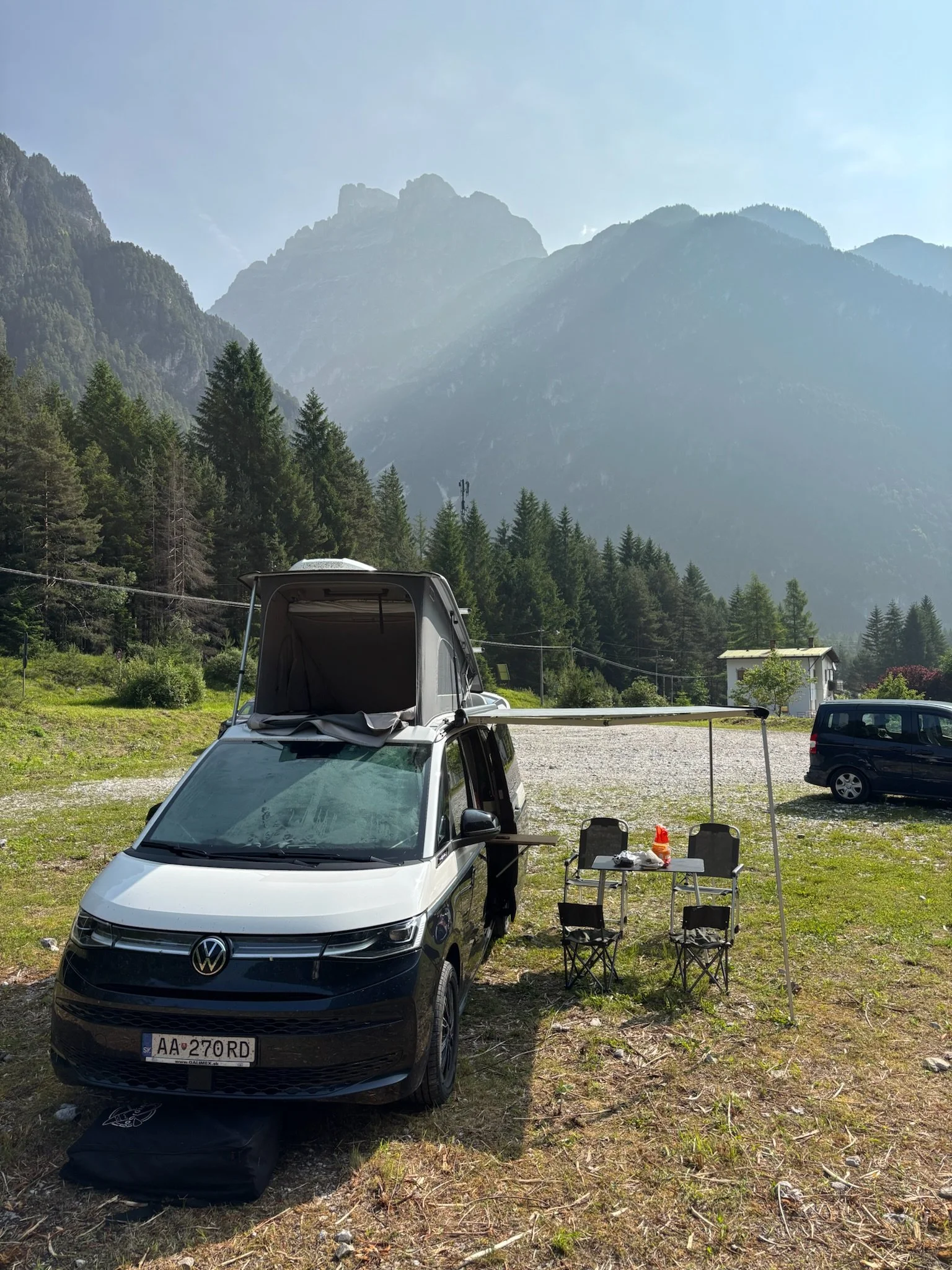A white van with a rooftop tent parked outdoors on a grassy area, with a small outdoor setup including a table and chairs, and mountains in the background.