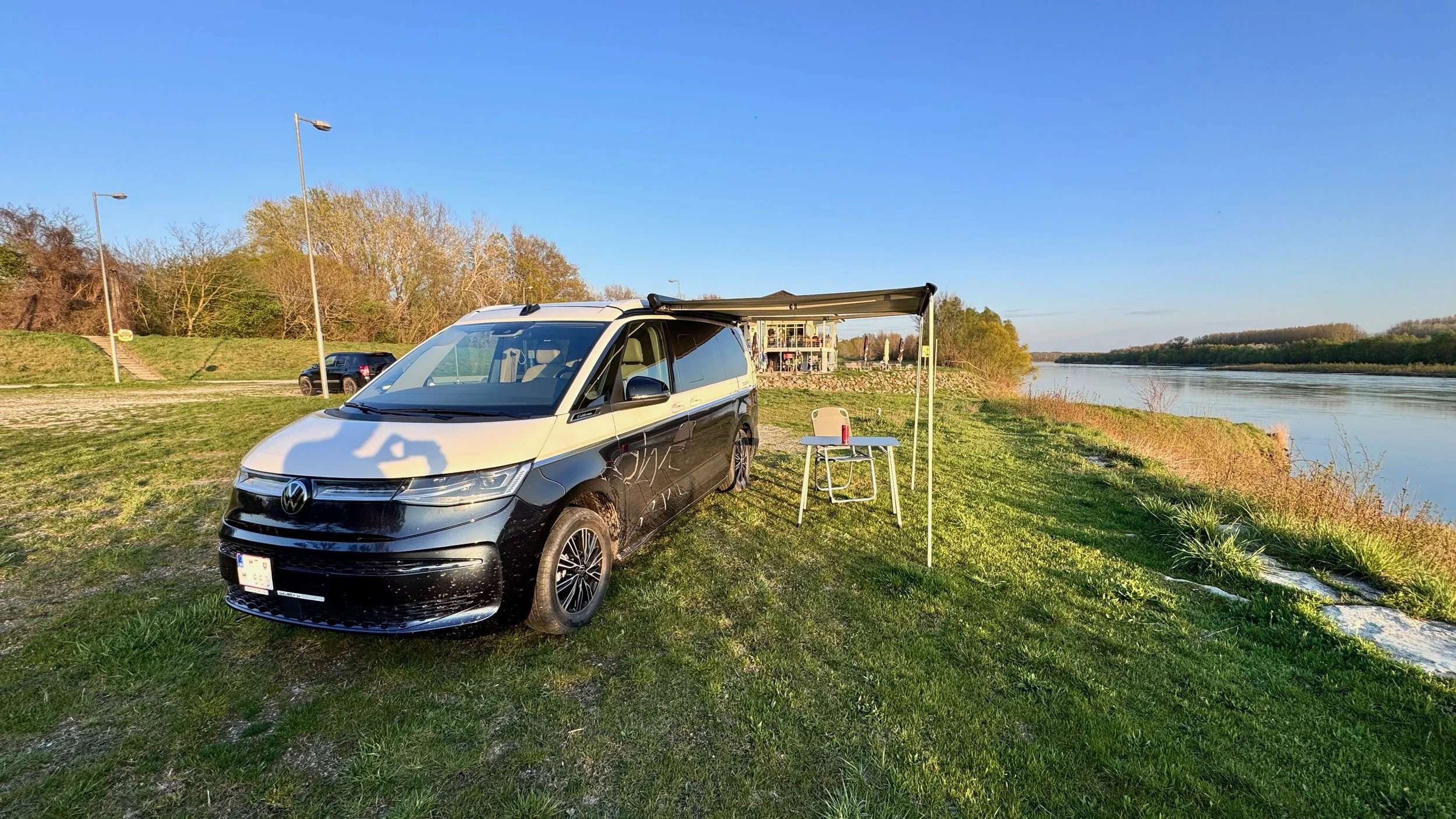 A black and white Volkswagen camper van parked on a grassy riverbank with a table, chair, and a small awning, next to a calm river with trees on the opposite bank and a blue sky overhead.