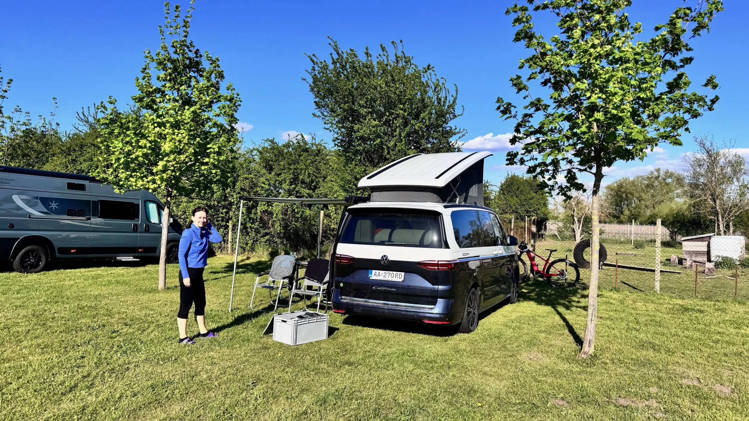 A person in a blue jacket standing next to a black Volkswagen camper van with a pop-up roof, parked on a grassy area with trees and other vehicles in the background.