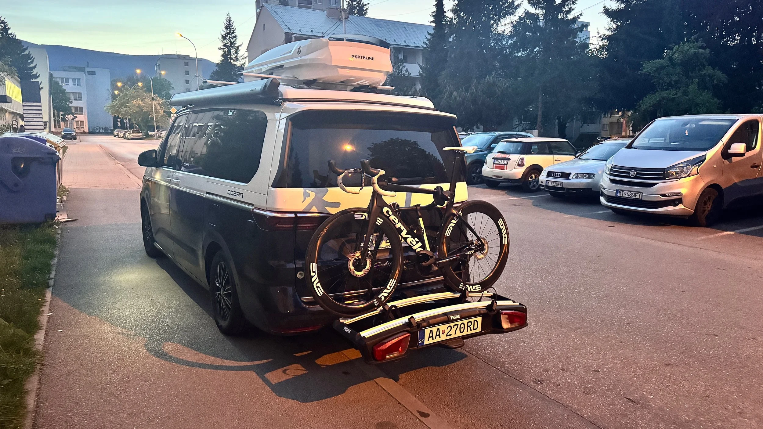 A black van with a roof cargo box and a bicycle mounted on a rear rack parked in a city street at dusk. Several other cars and buildings are visible in the background.