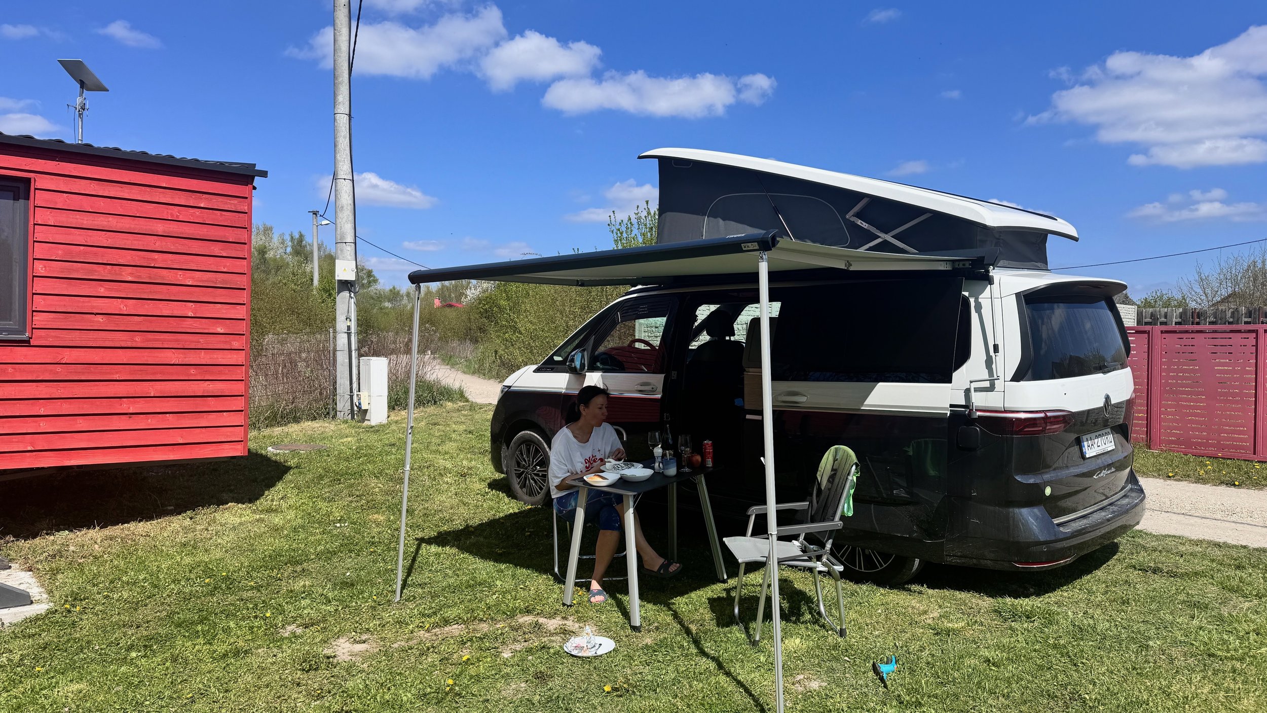 A woman sitting at a small outdoor table with food and drinks next to a black and white camper van with a raised roof, on a grassy area under a blue sky with scattered clouds. There's a red house with a window and a utility pole in the background.
