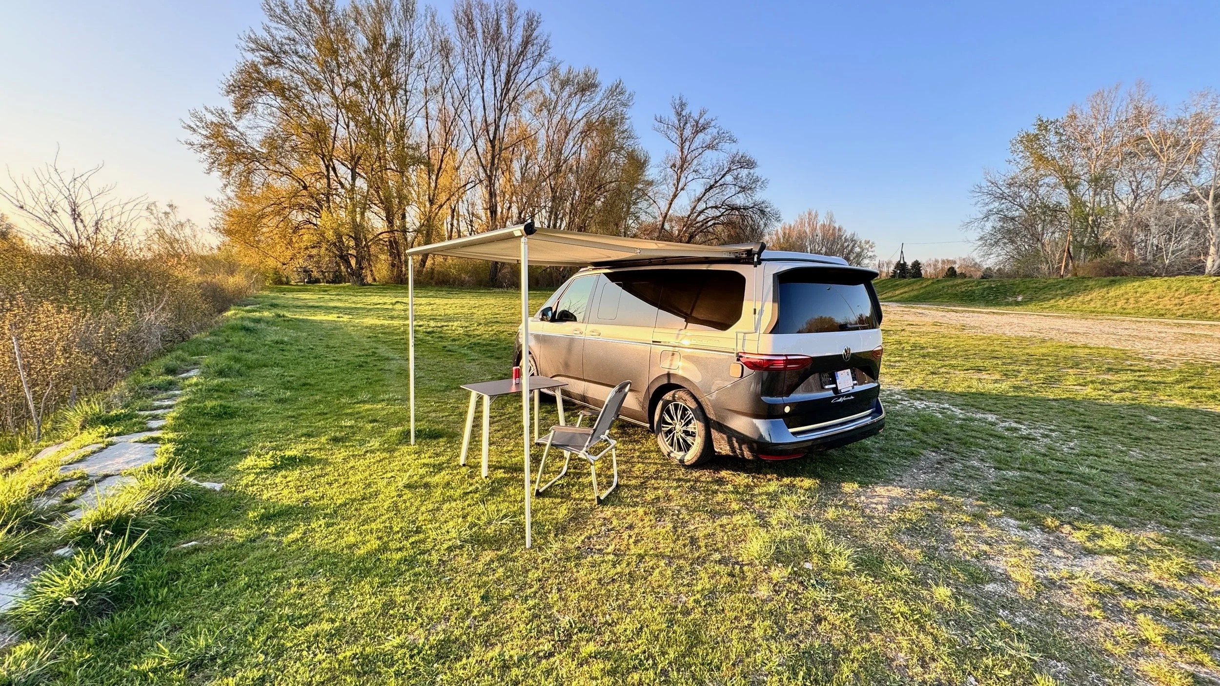 A van parked on a grassy area with a canopy attached to it, a small table, and a chair, with trees and a dirt path in the background during sunset.