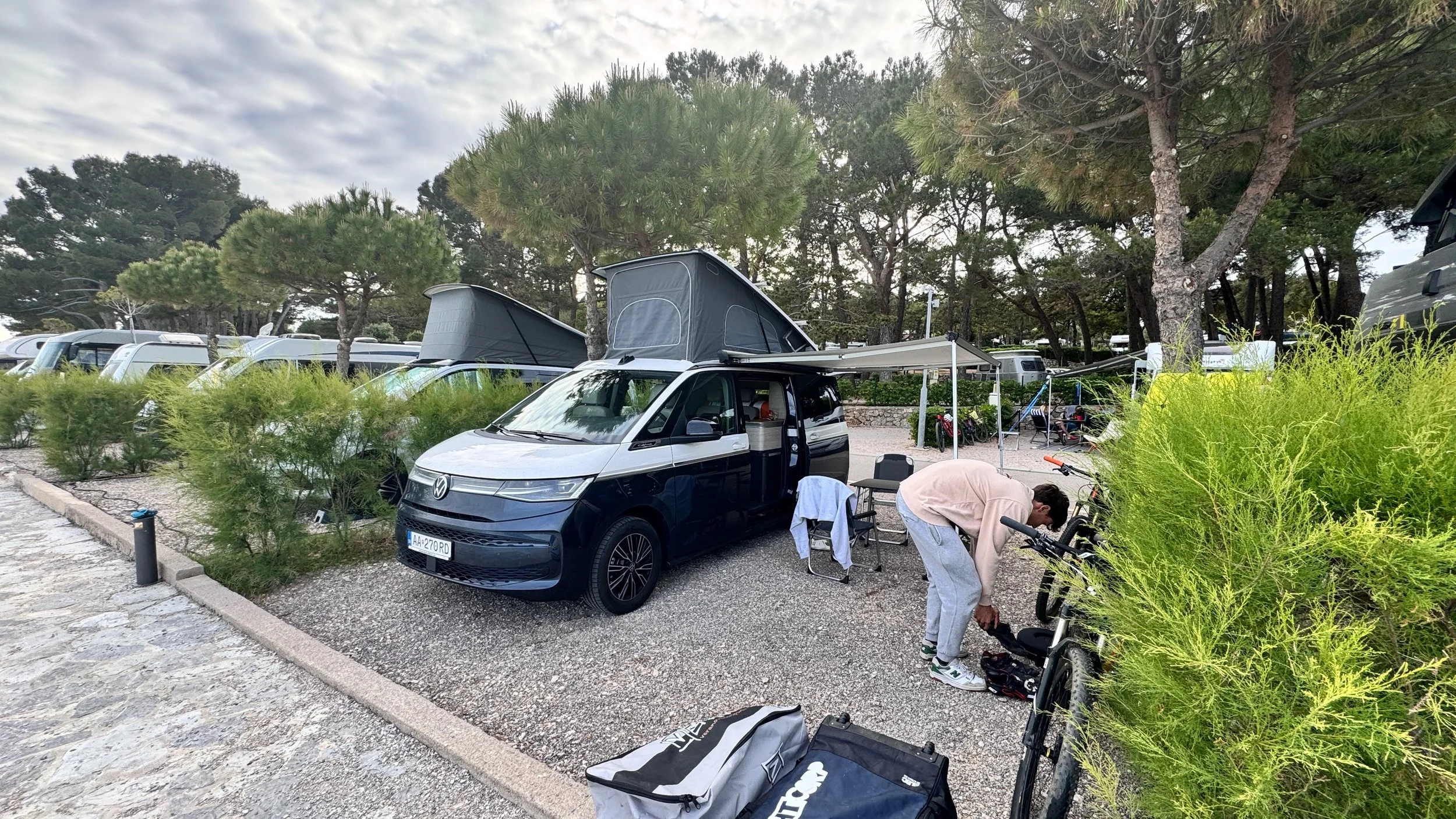 A person preparing a bicycle next to a Volkswagen camper van with a pop-up roof at a campground surrounded by trees and parked vehicles.