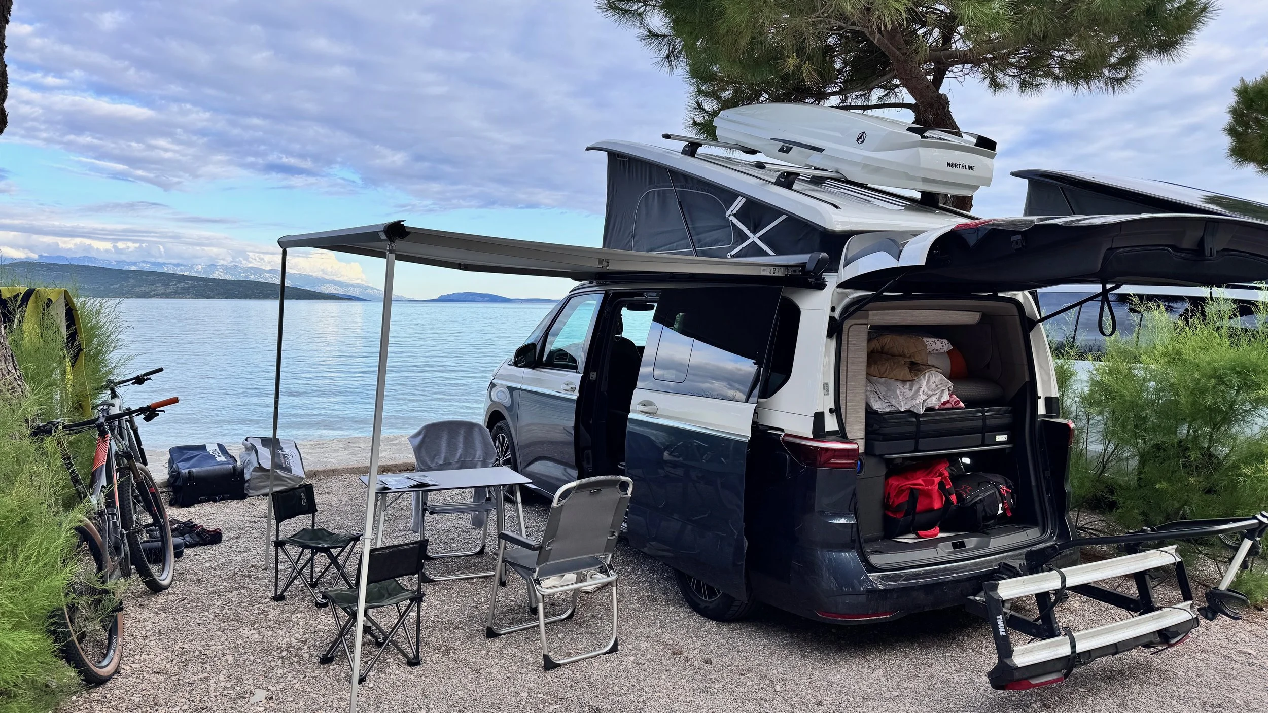 A camper van parked by a lakeside with camping equipment, bicycles, and outdoor furniture, overlooking a large body of water with distant land and a partly cloudy sky.
