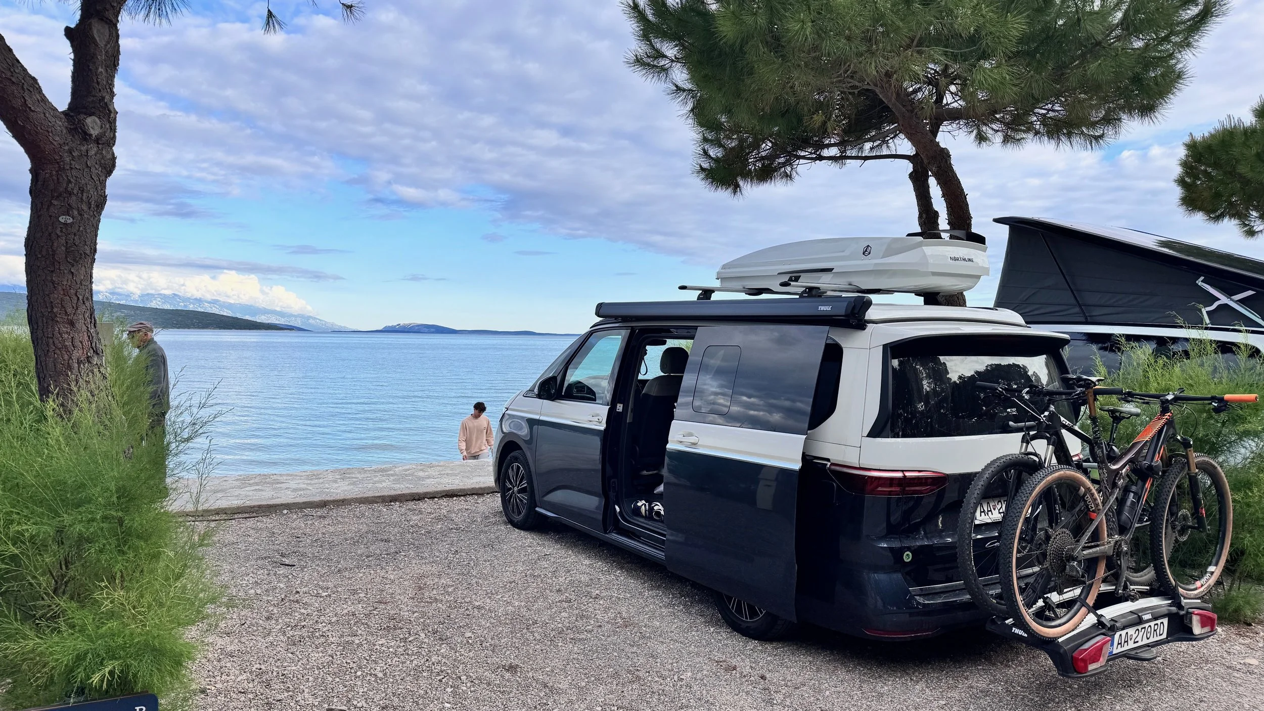 A black and white camper van parked near a lake with two bikes mounted on the back, surrounded by trees and gravel, with two people sitting near the water and a mountain in the distance under a partly cloudy sky.