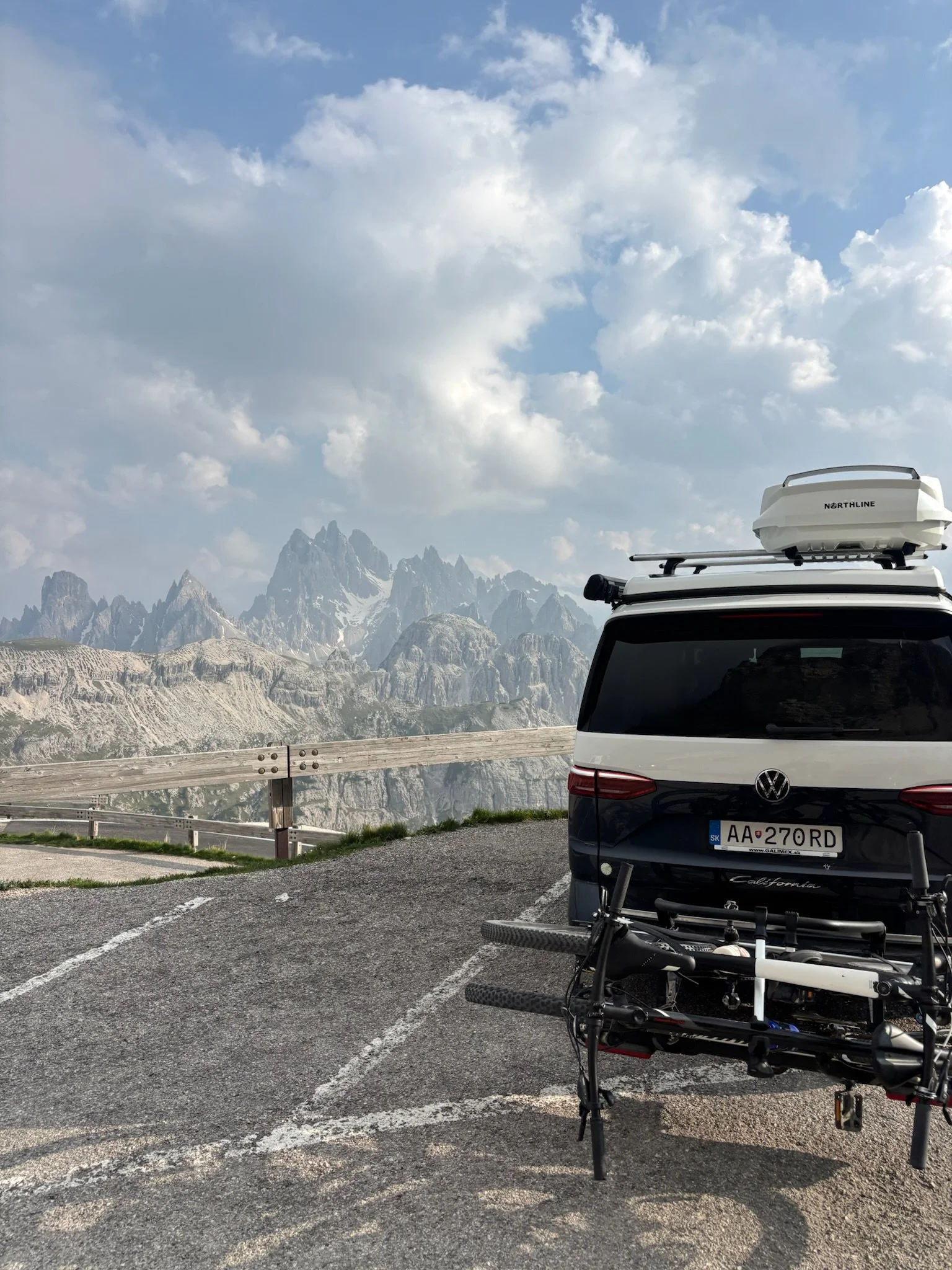 A Volkswagen SUV parked on a mountain overlook with a bicycle mounted on the back, overlooking rugged mountain peaks and a partly cloudy sky.