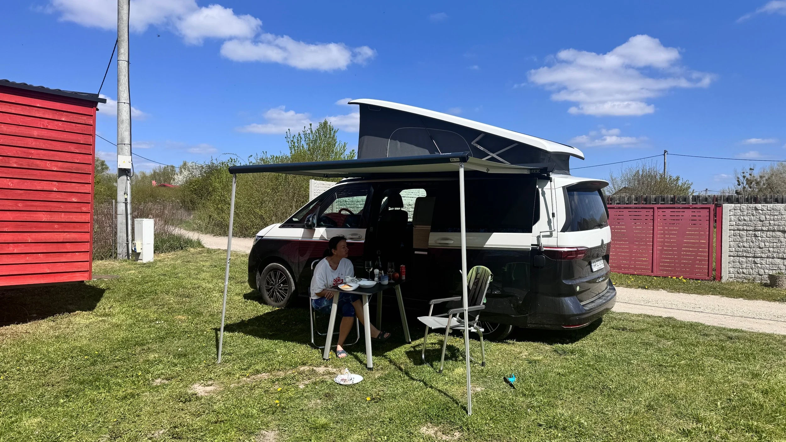 A person sitting at a small table outside a camper van, with a black and white exterior, parked on grass near a red fence and greenery, under a blue sky with scattered clouds.