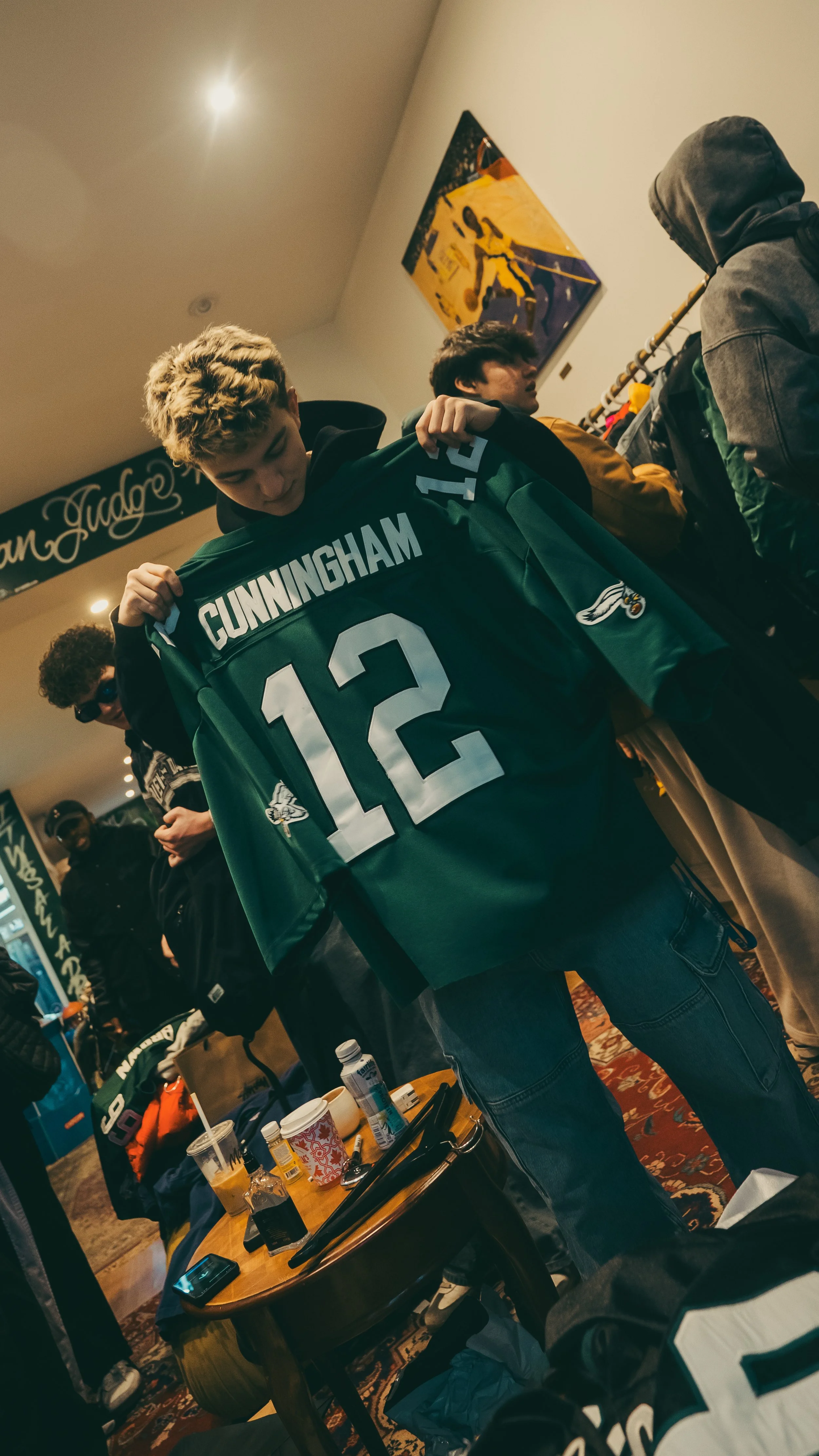A young man holding a green Philadelphia Eagles football jersey with the name Cunningham and the number 12, surrounded by others in a room with sports memorabilia and jackets.