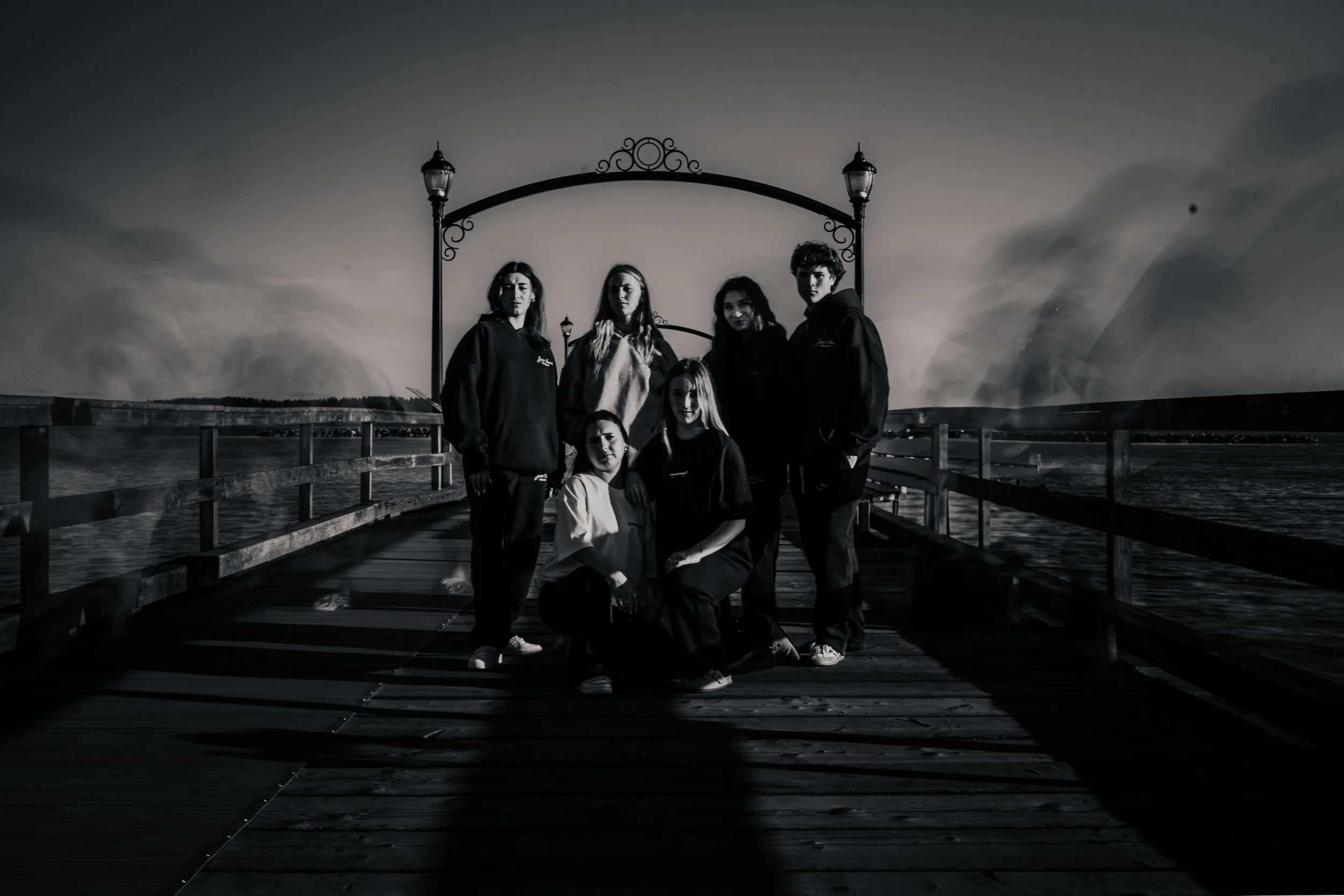 A group of six women standing and kneeling on a wooden dock near water, with a decorative arch and street lamps in the background, captured in black and white.