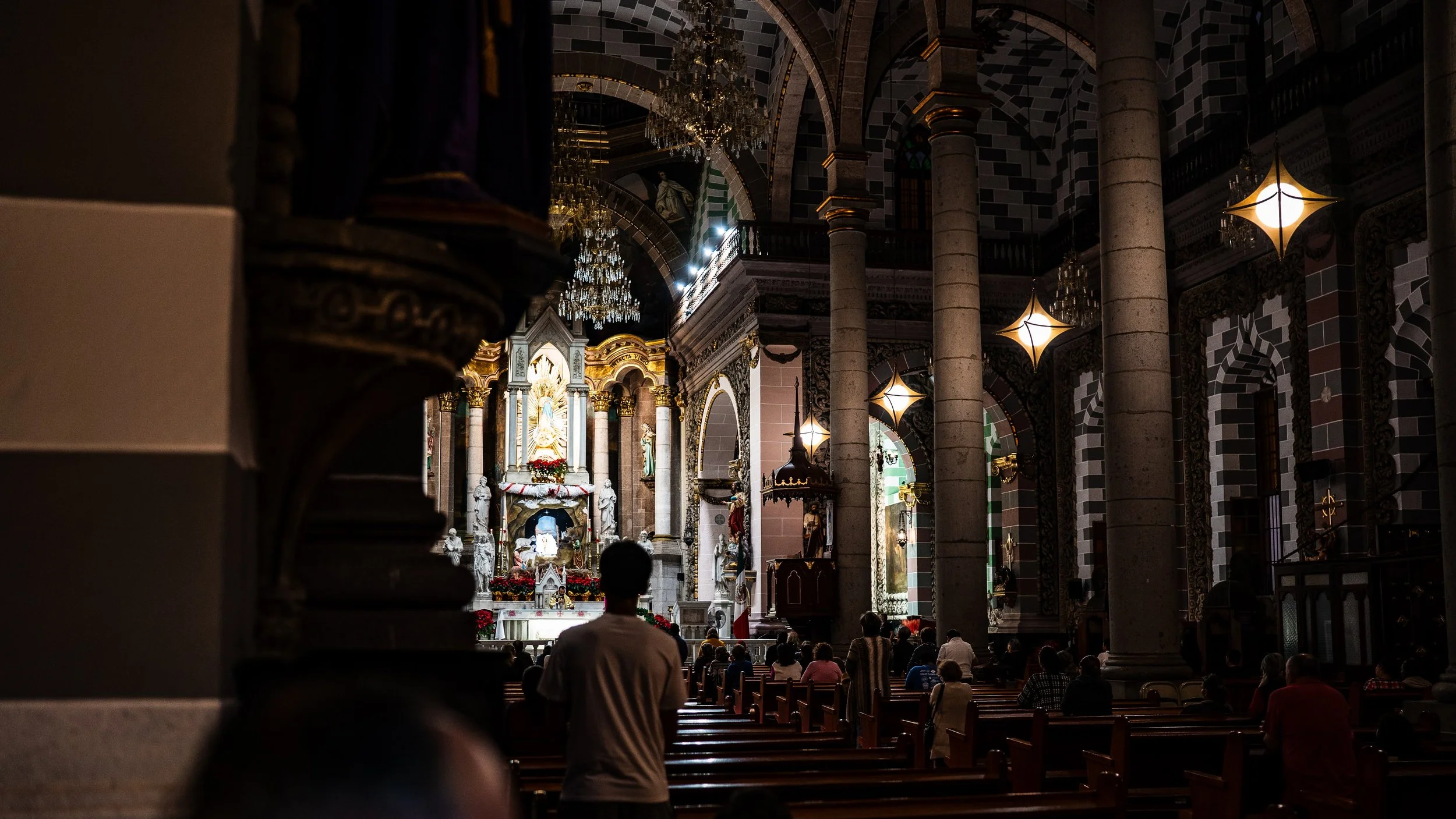 Interior of a church with tall columns, stained glass windows, and ornate decorations, with people seated in pews and attending a service or prayer.