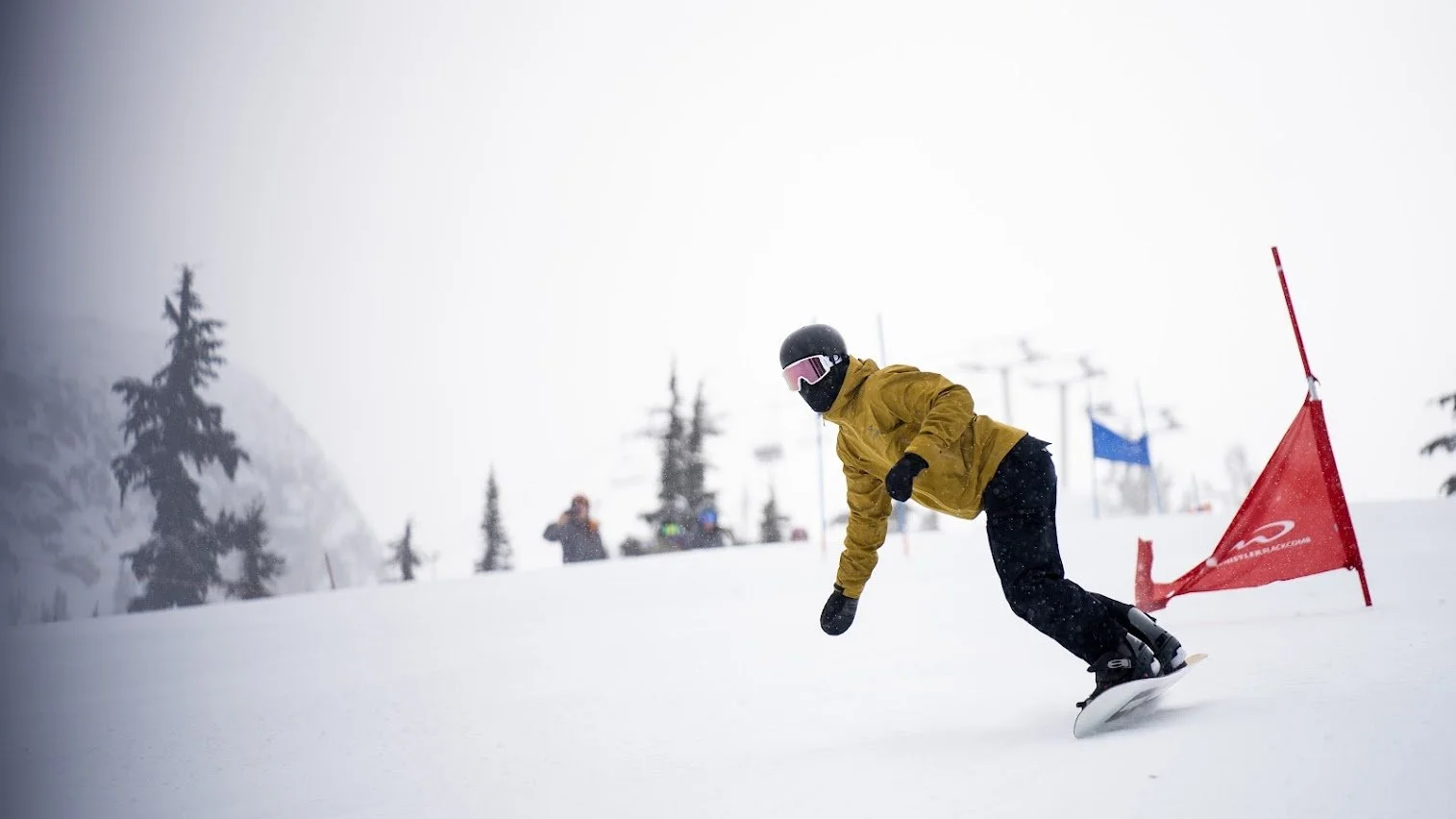 A person snowboarding downhill on a snowy mountain slope, wearing a yellow jacket, black helmet, and pink-tinted goggles, with a red slalom gate nearby.