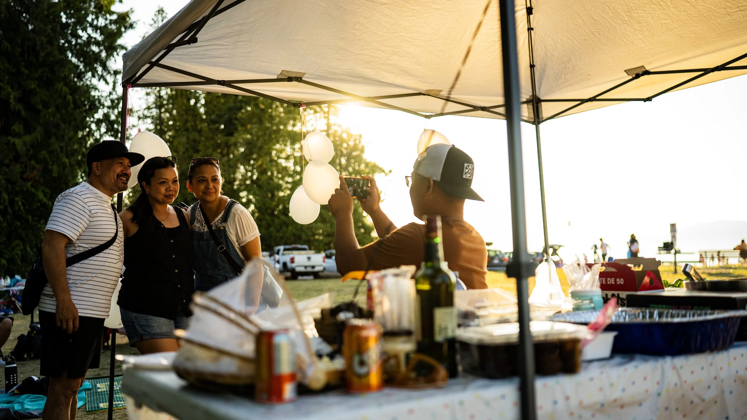 Four people standing under a tent outdoors, taking a photo of a group of three women. The scene is backlit by the setting sun, with trees and parked cars in the background, and a table with food and drinks in the foreground.