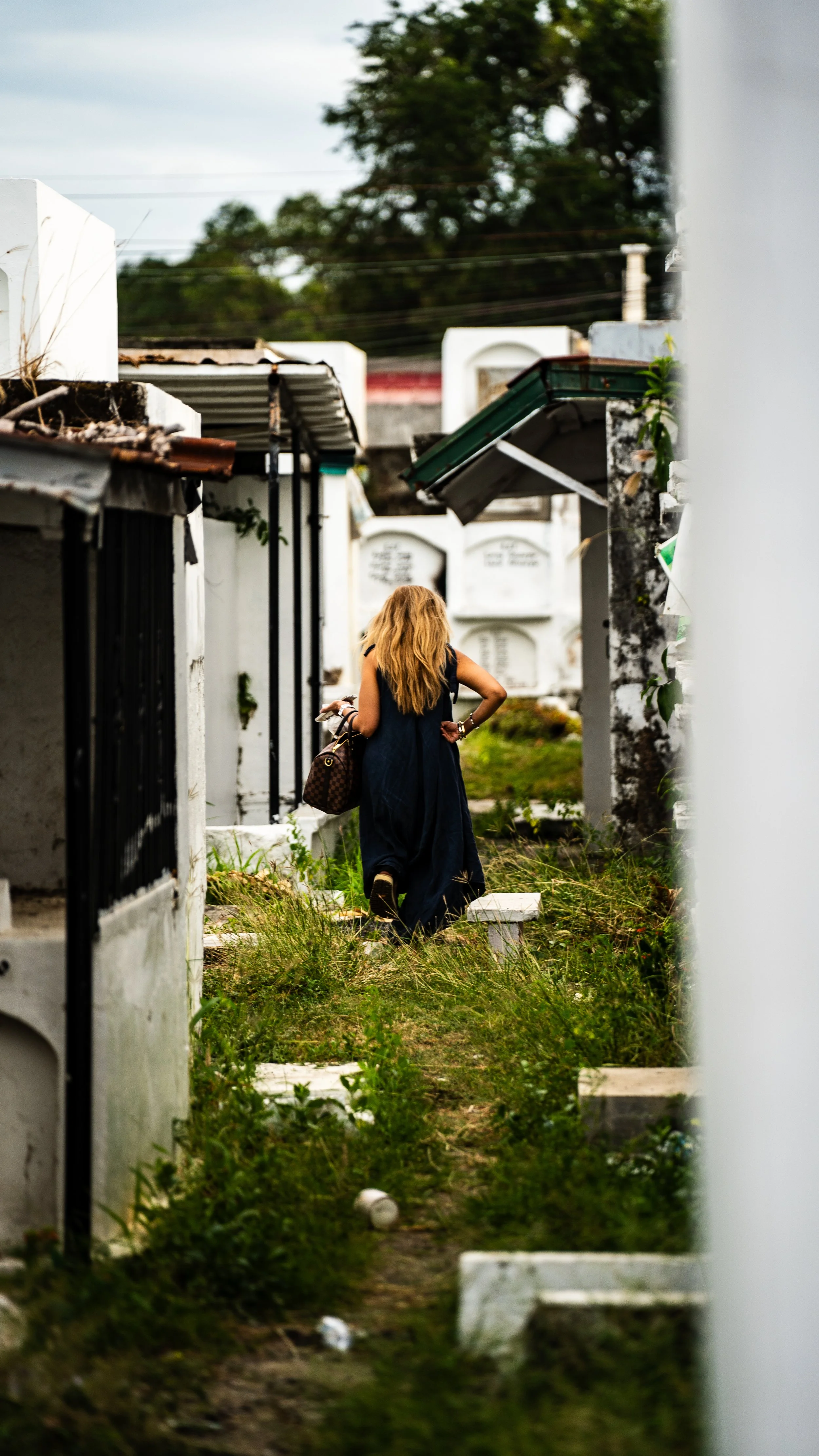 A woman with long, wavy blonde hair wearing a dark blue dress, carrying a patterned handbag, walking through a cemetery with white tombstones and overgrown grass.