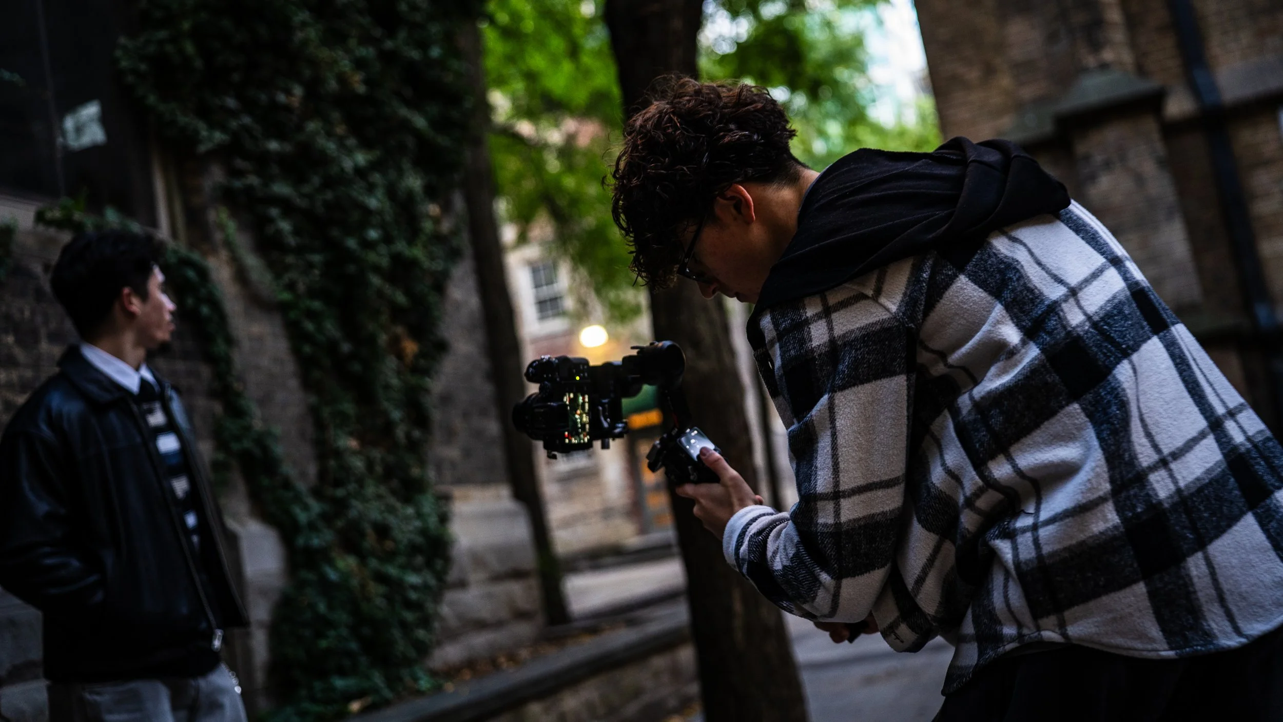 A person with curly hair and glasses is filming or taking a photo of a young man standing against a brick wall with ivy and trees in the background, on a city street during evening.