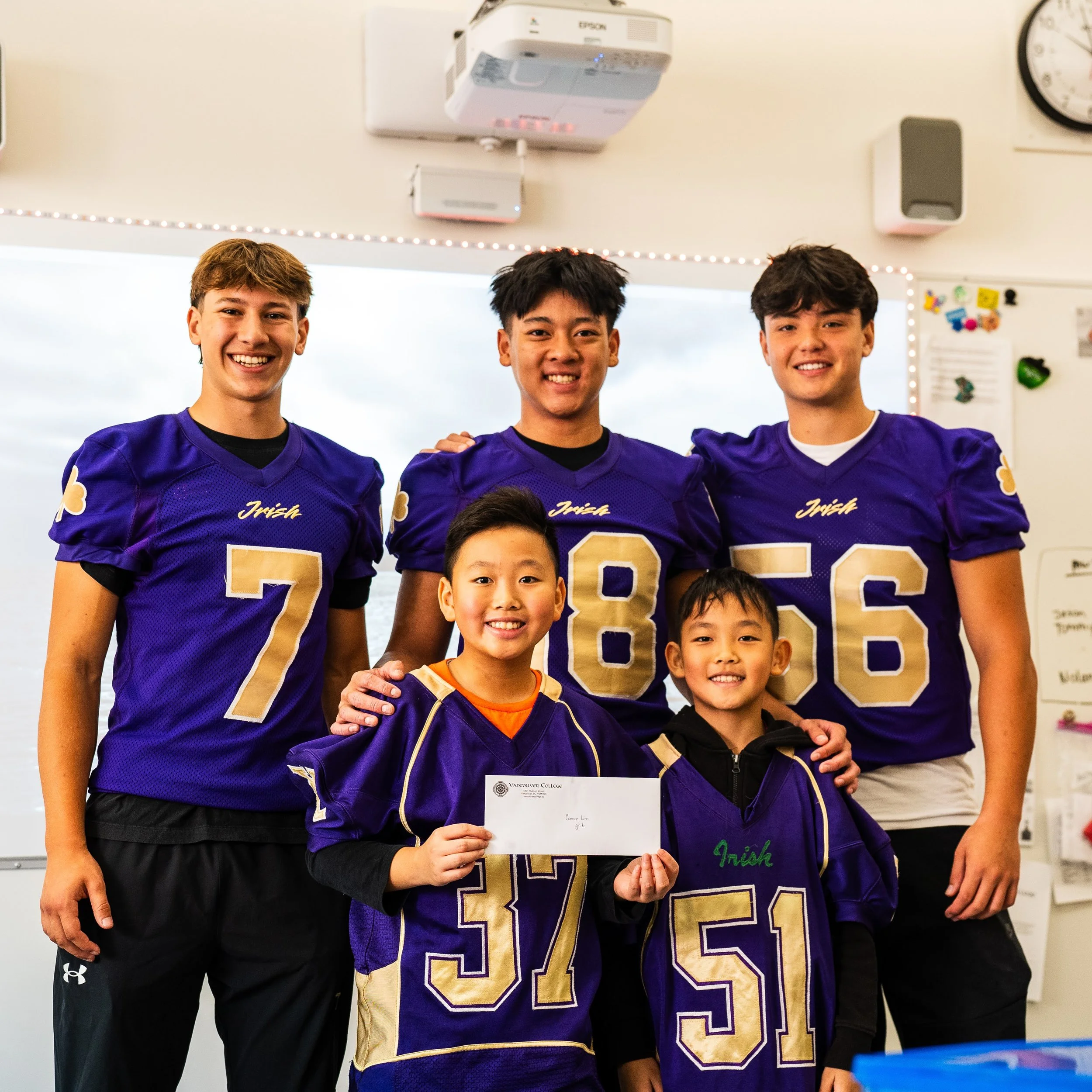 Five young males, three wearing purple football jerseys with gold numbers, smiling in a classroom, with two younger boys holding a check and a jersey.
