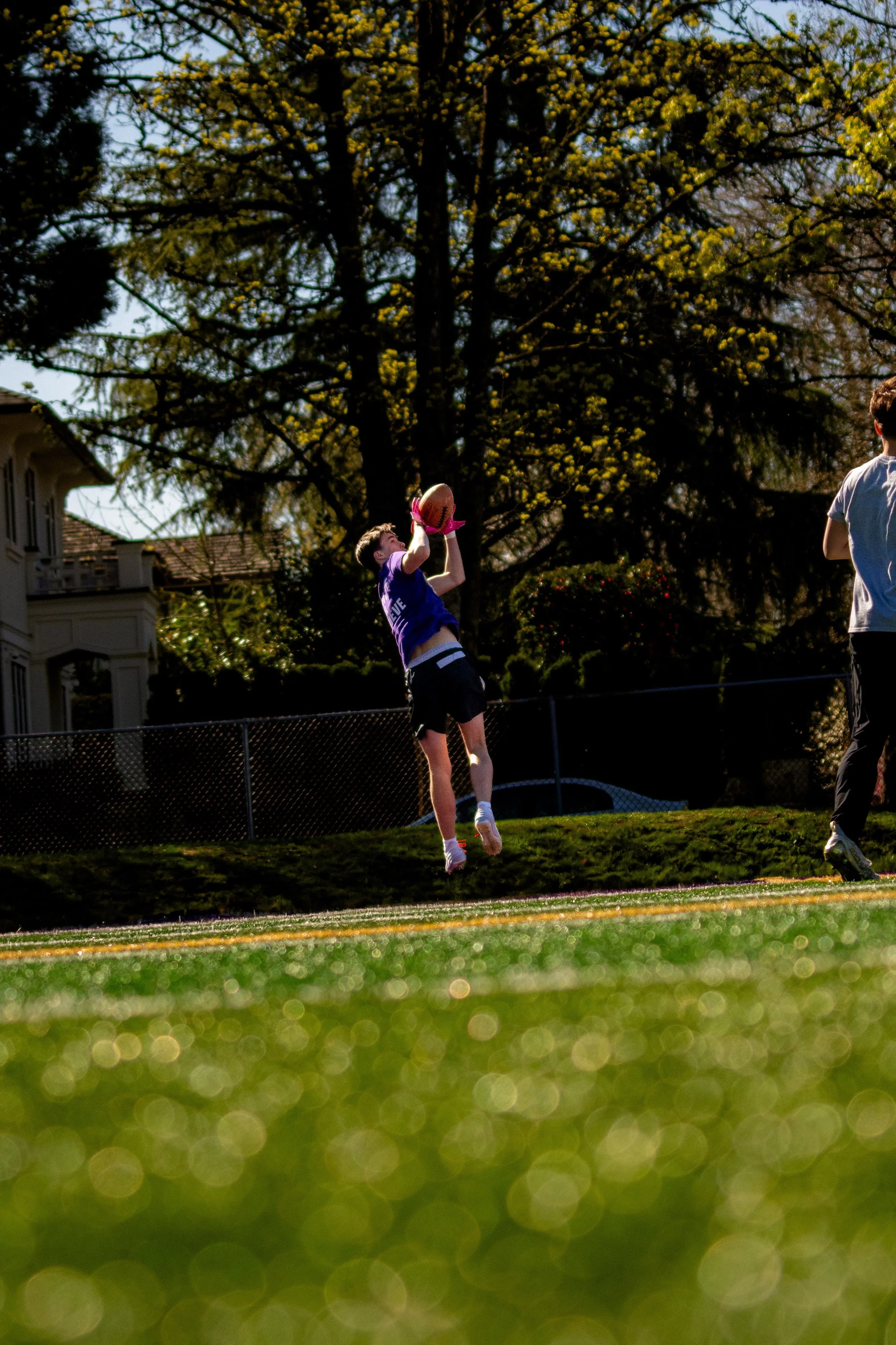 A young person jumping to catch a football on a sports field during daytime, with a large tree and houses in the background.