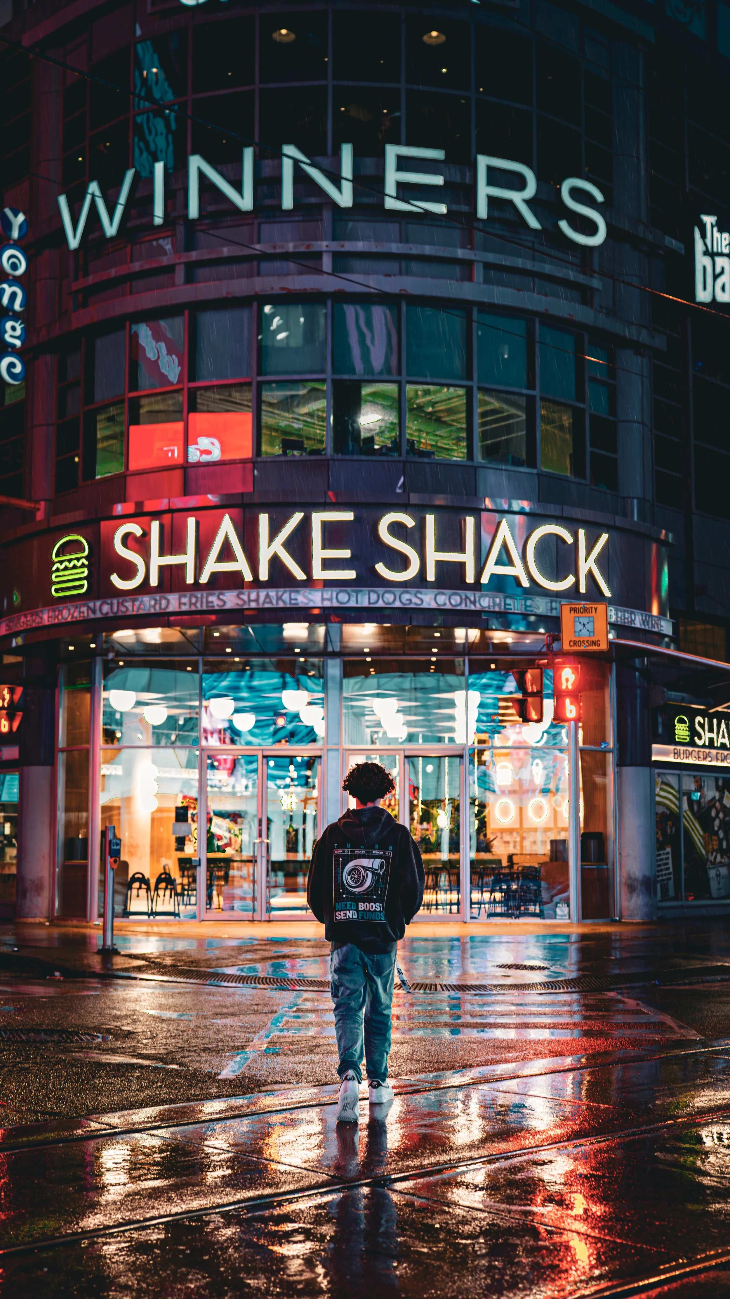 A person standing outside a Shake Shack restaurant in an urban area at night, with neon signs and reflections on the wet pavement.