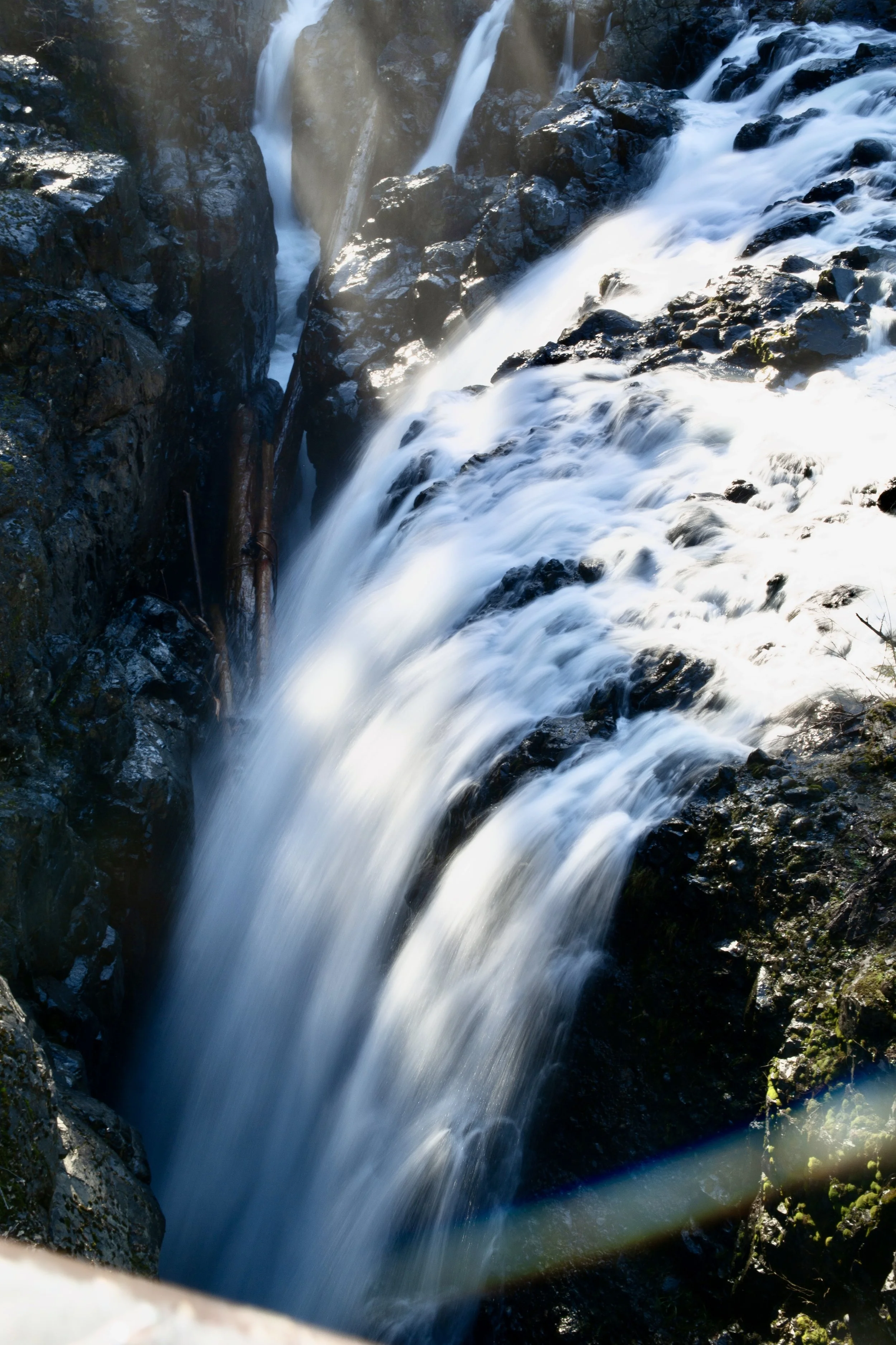 Waterfall cascading over dark rocks in a forested area with sunlight filtering through.