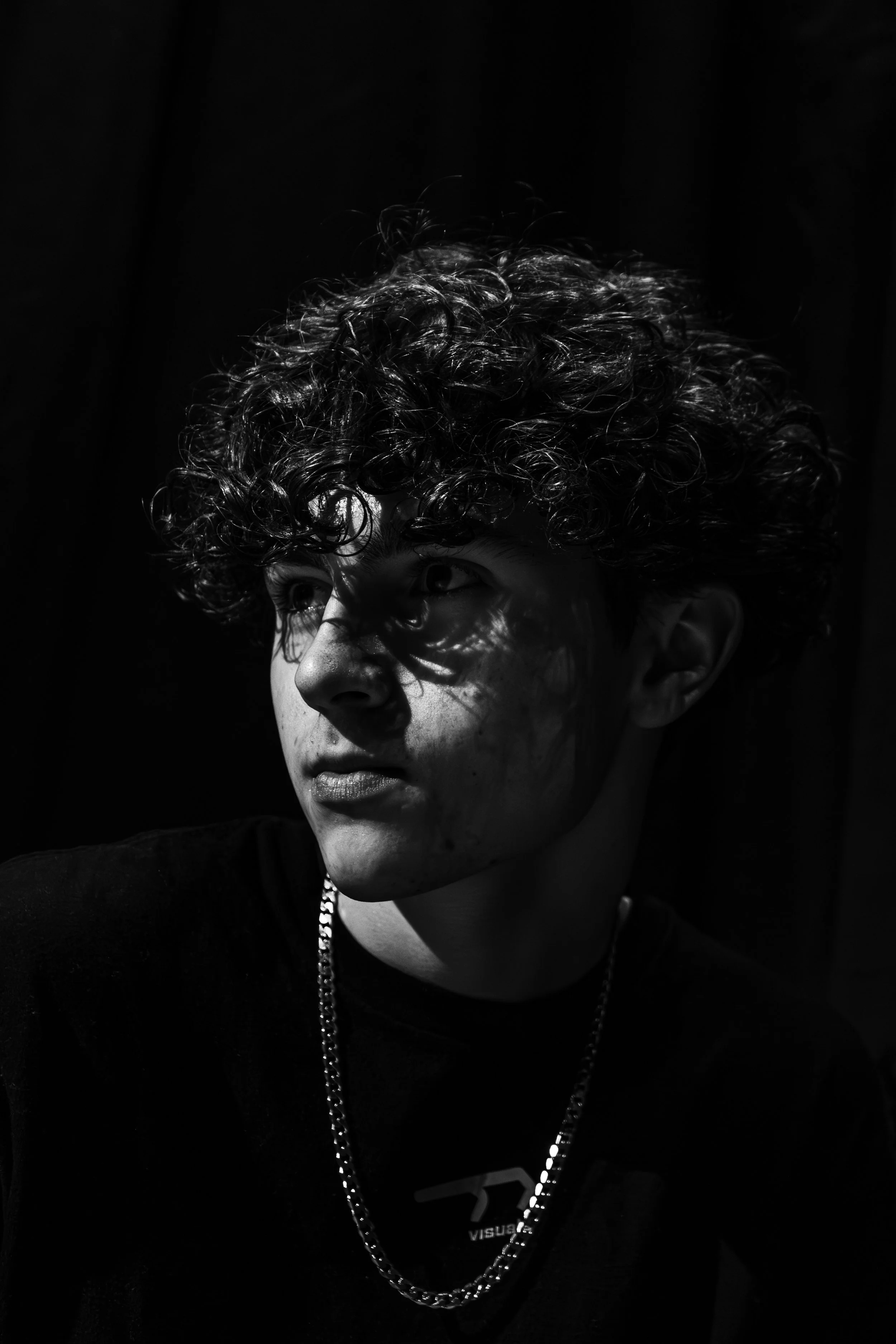 Black and white portrait of a young person with curly hair and a chain necklace, with shadows cast across their face in a low-light setting.