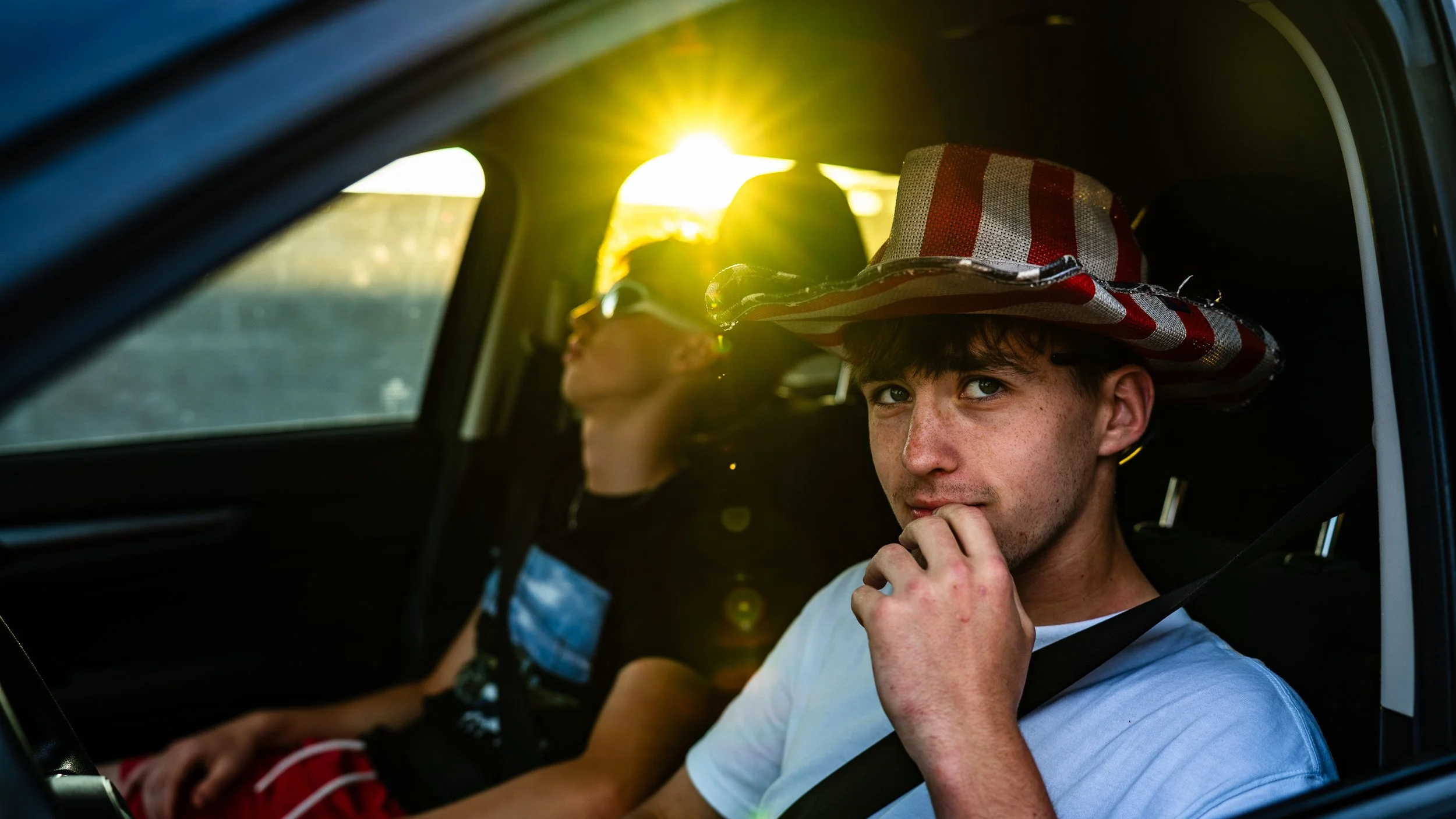 Young man wearing a wide-brimmed striped hat sitting in a car seat, looking at the camera with a thoughtful expression, with a woman in the background wearing sunglasses, both in a vehicle during sunset.