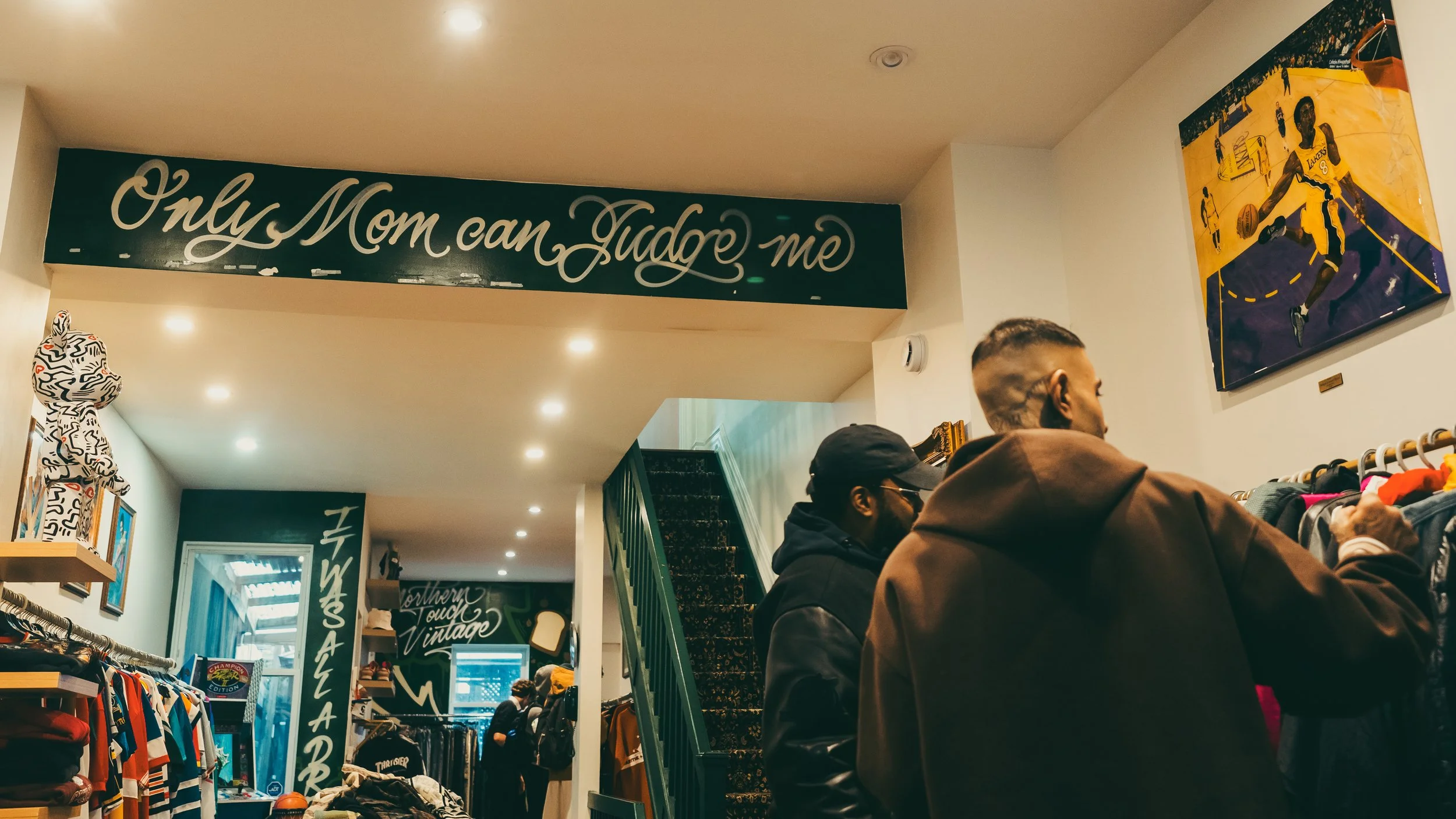 Inside a vintage clothing store with shoppers browsing. A large sign says 'Only Mom can Judge me.' Wall art features a basketball player in action.