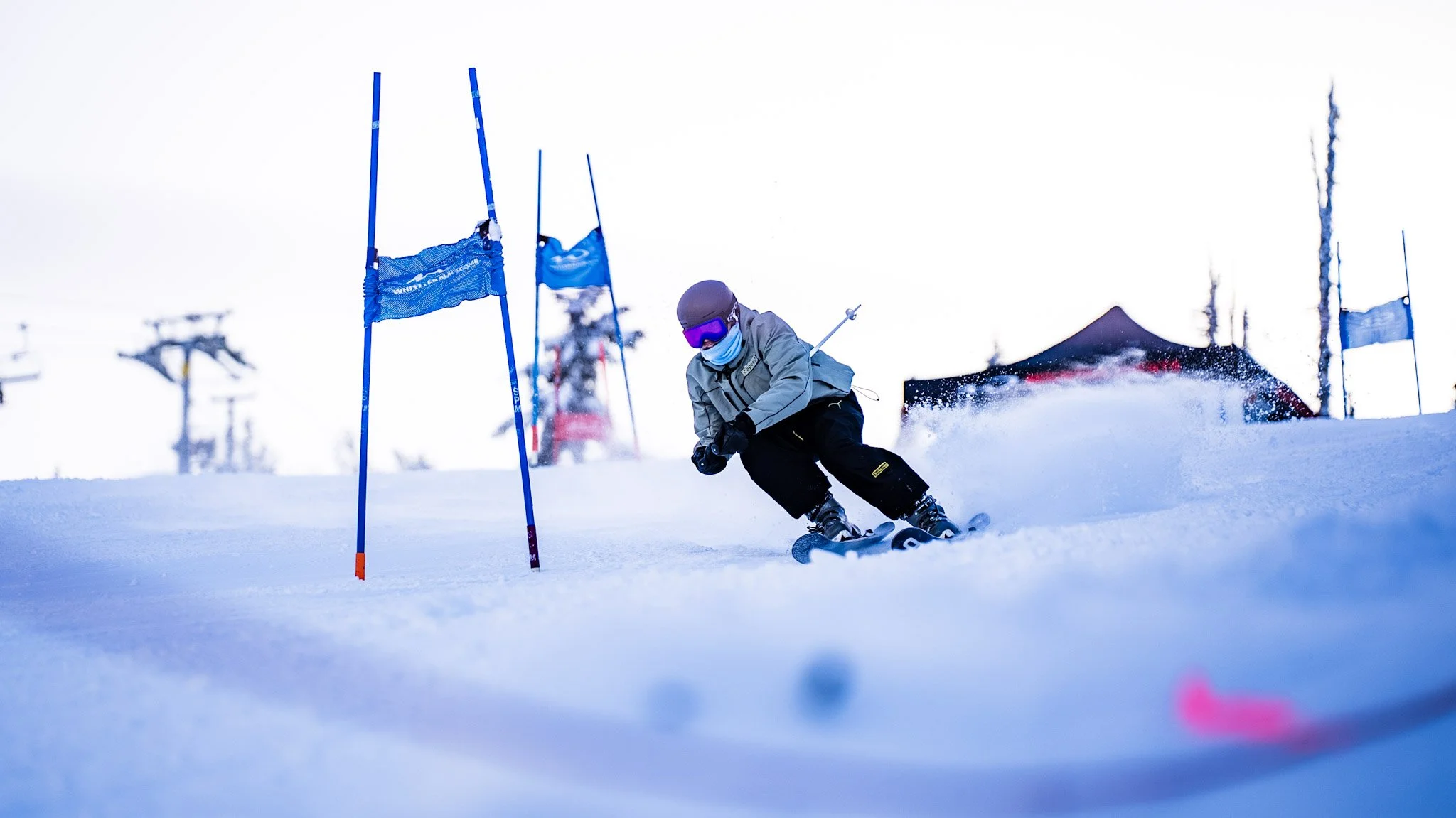 A young skier wearing a gray jacket, black pants, purple goggles, and a face mask takes a sharp turn on a snowy slope during a ski race, passing between blue slalom gates with a tent and ski lifts visible in the background.