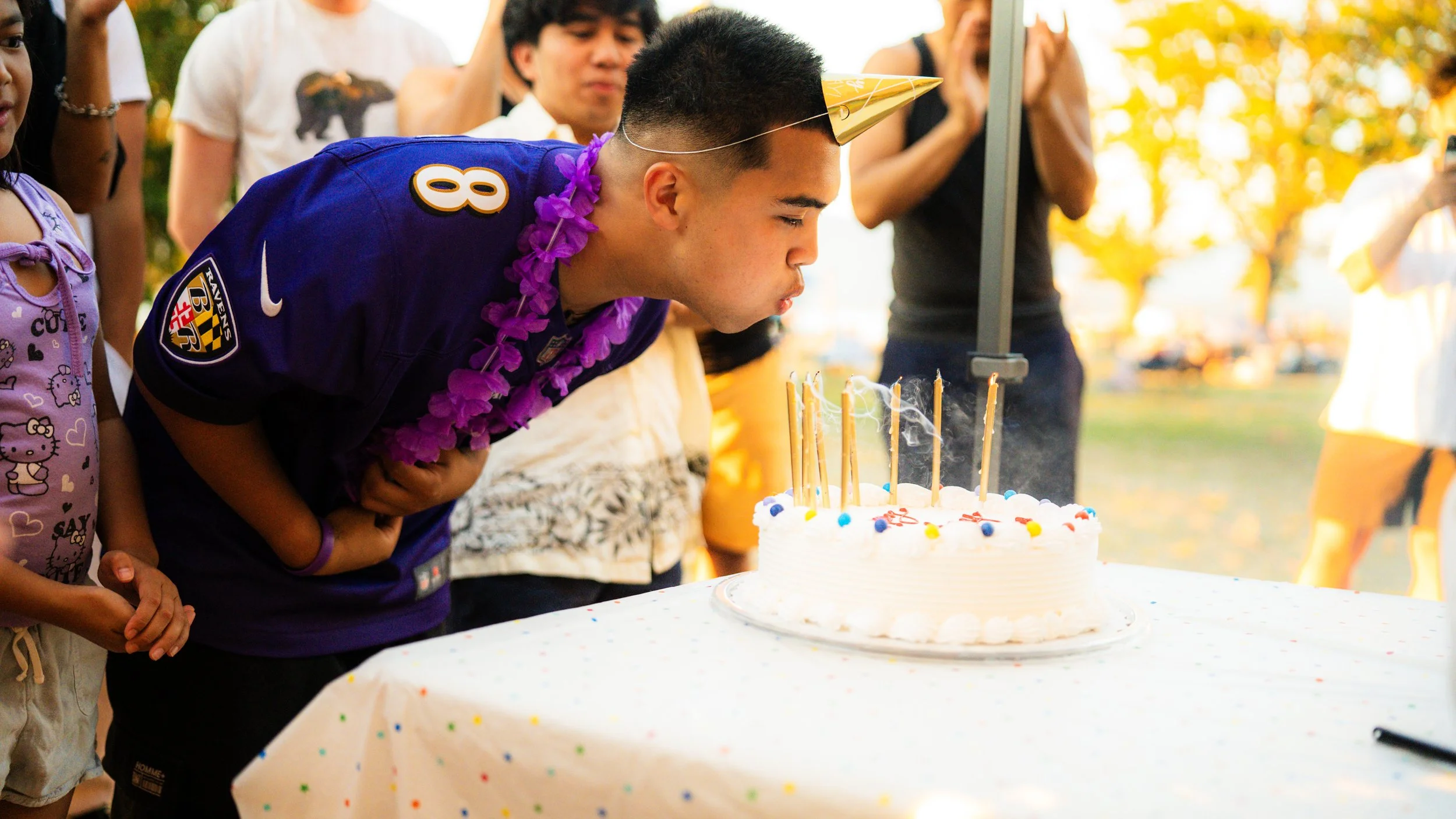 A young man in a purple sports jersey and party hat blows out candles on a birthday cake with other people watching outdoors during sunset.