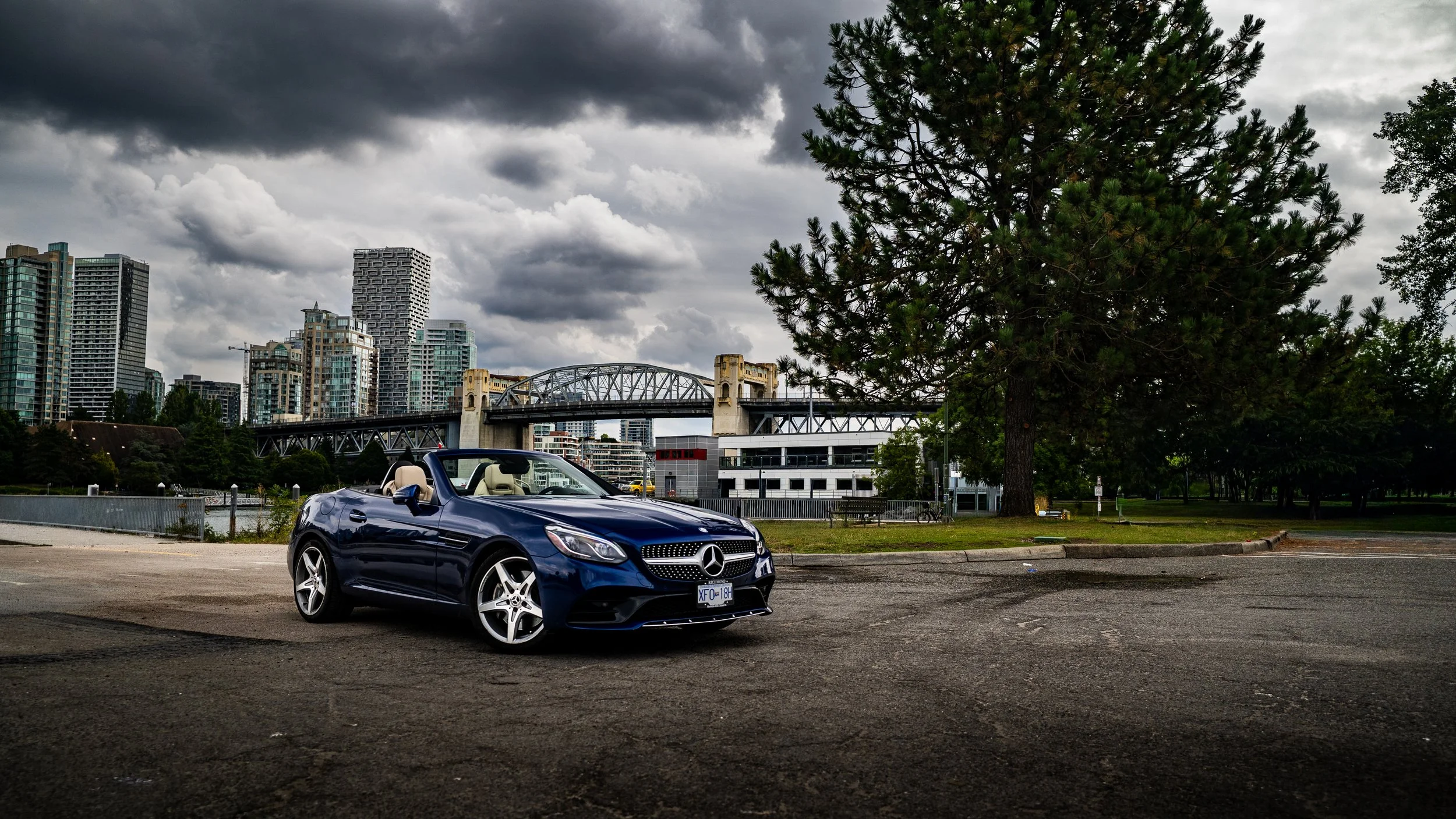 A blue Mercedes-Benz convertible car parked in an urban lot with a city skyline and a large tree in the background, under a cloudy sky.