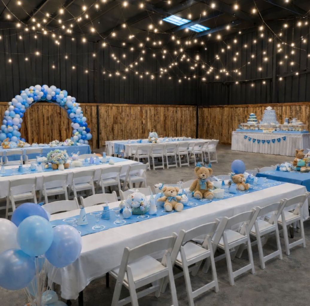 Decorated indoor celebration venue with blue and white theme, teddy bears, balloon arrangements, table settings, and a cake table.