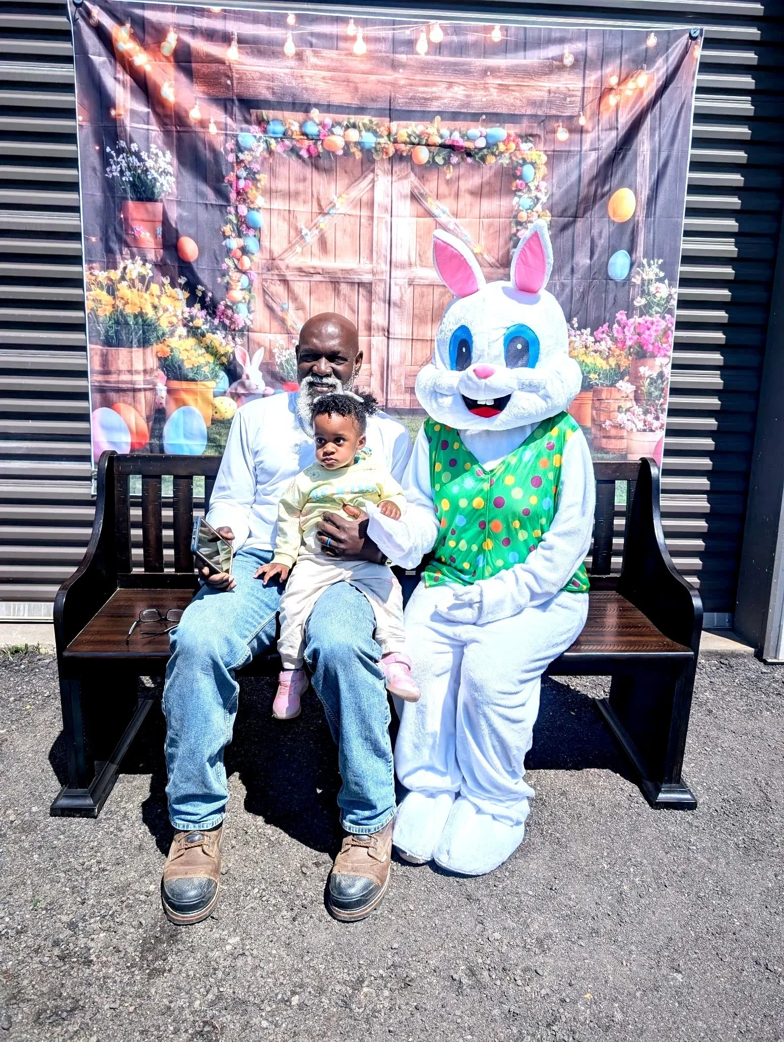 Man, child, and person in Easter Bunny costume sitting on a bench outdoors with Easter-themed backdrop.
