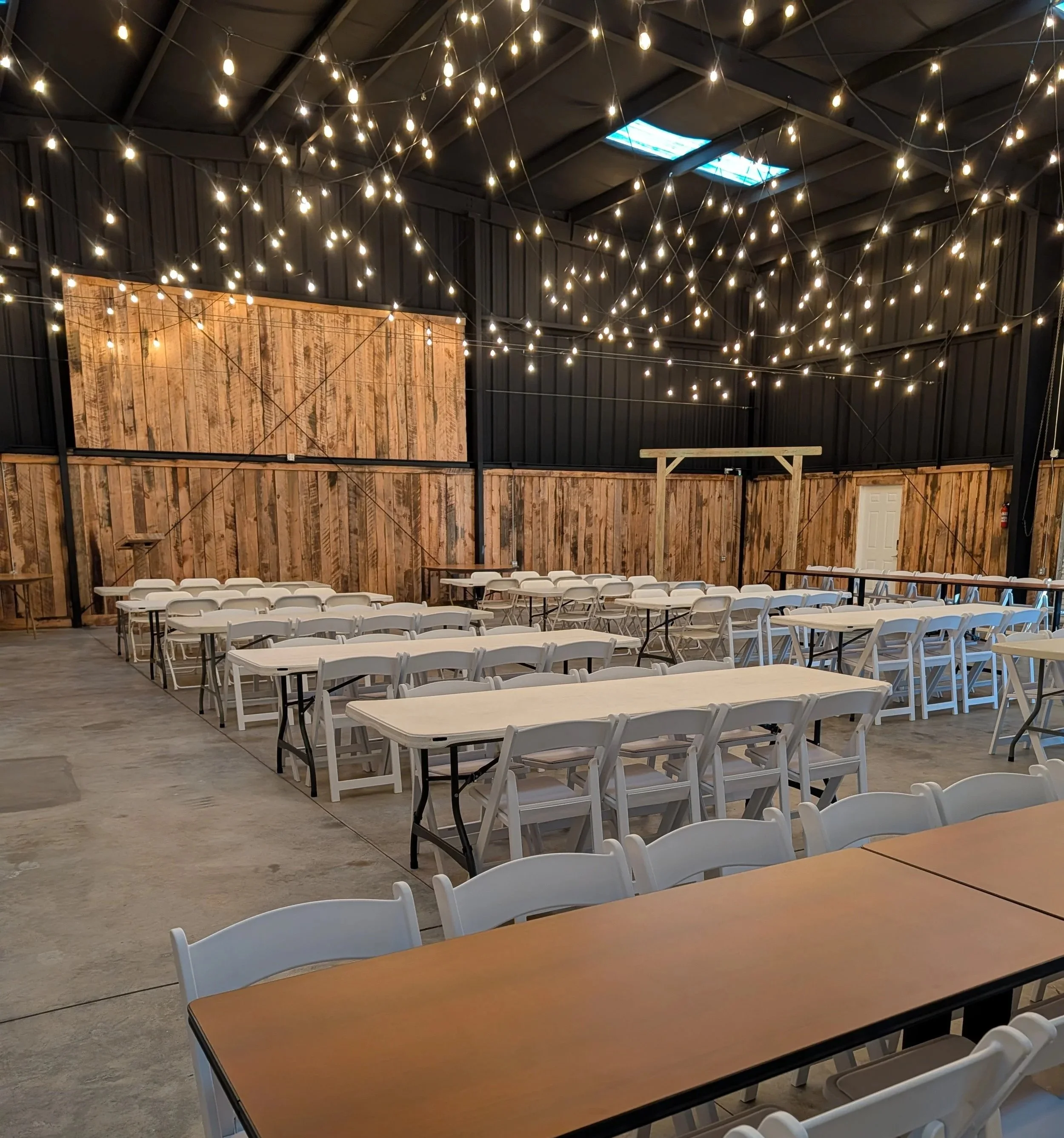 An indoor event space with string lights hanging from the ceiling, wooden wall panels, and multiple tables and chairs arranged for a gathering.