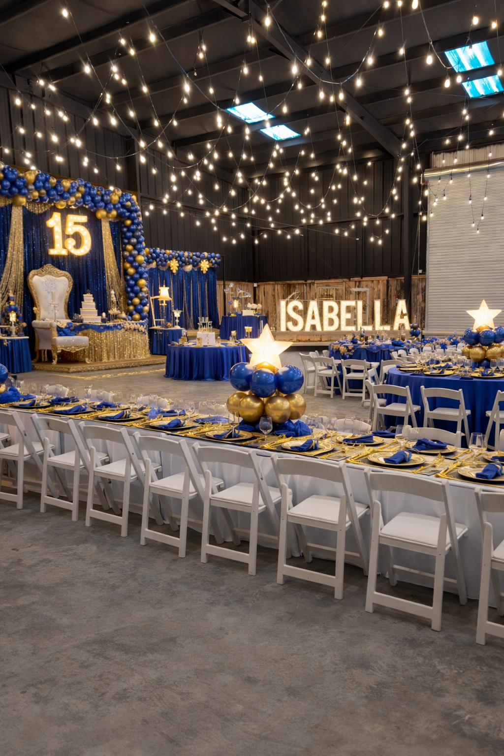 Decorated celebration hall with blue, gold, and white theme, featuring string lights, table settings, balloons, and a large illuminated name sign 'SABELLA' and a number 15 sign indicating a quinceañera celebration.