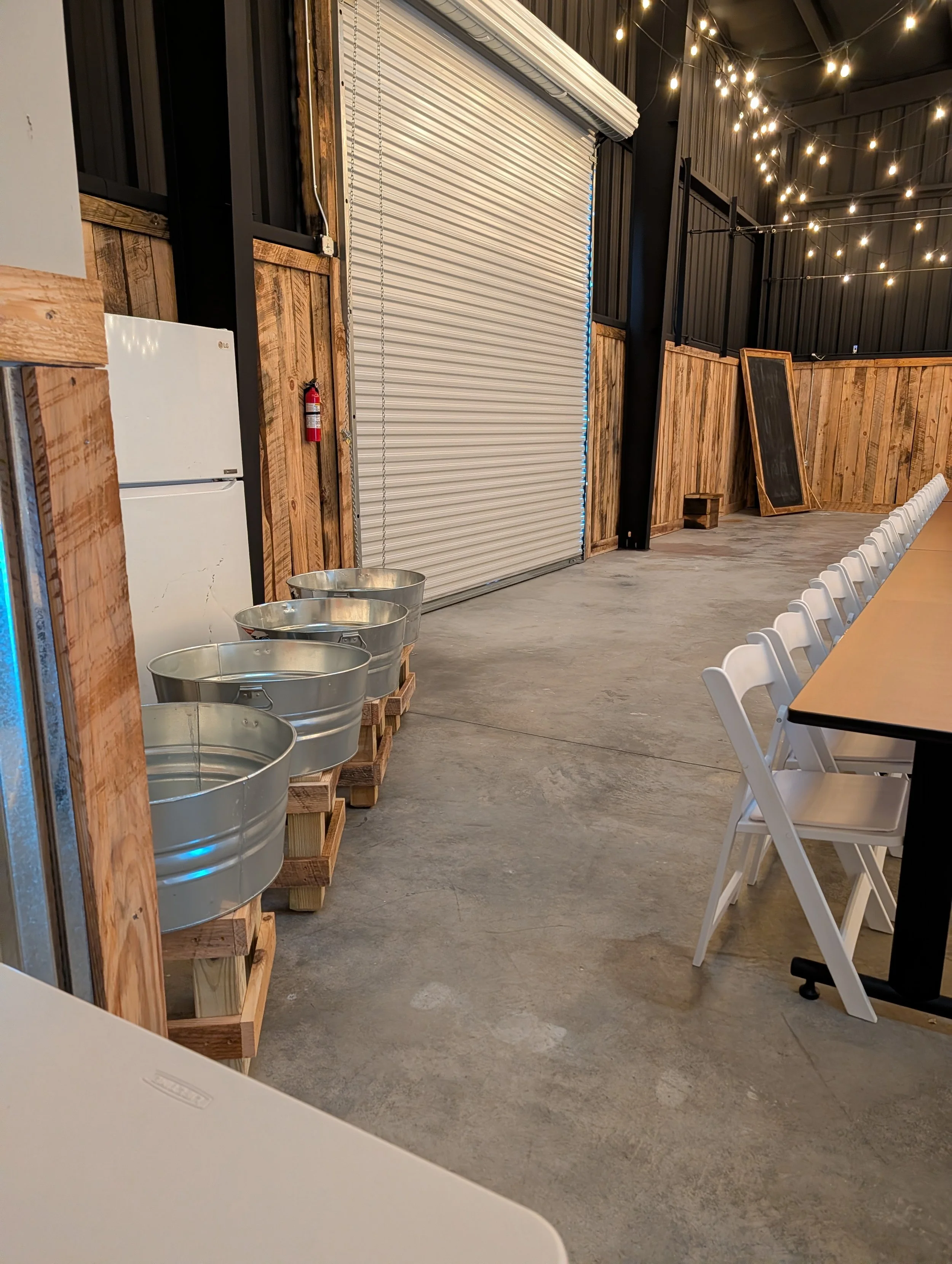 Indoor event space with string lights hanging from the ceiling, metal buckets on wooden pallets, a white refrigerator, and a large metal roll-up door. There are white folding chairs along a long table and a chalkboard leaning against a wooden wall.