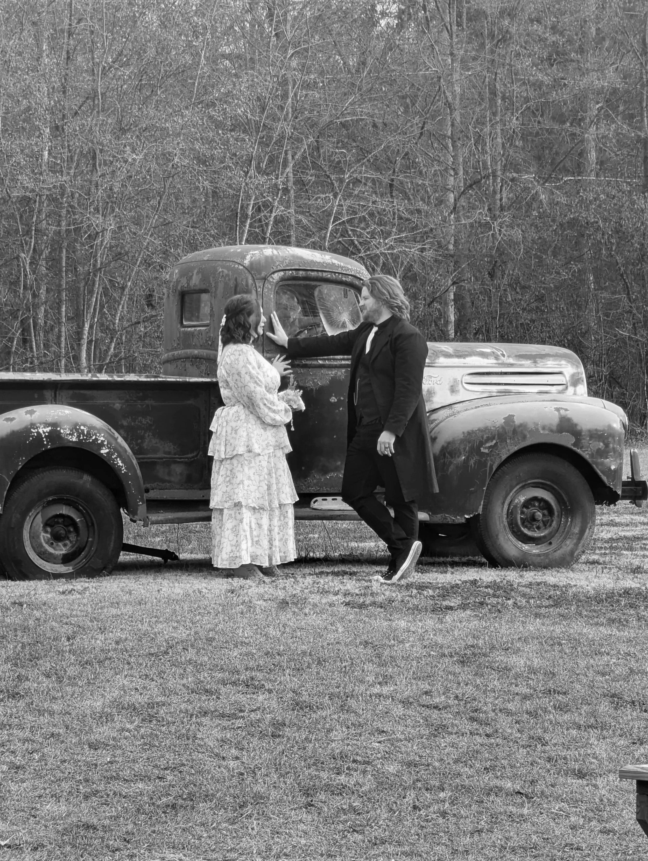A man and woman in vintage clothing sharing an intimate moment beside an old rusted pickup truck in an outdoor setting with trees in the background.