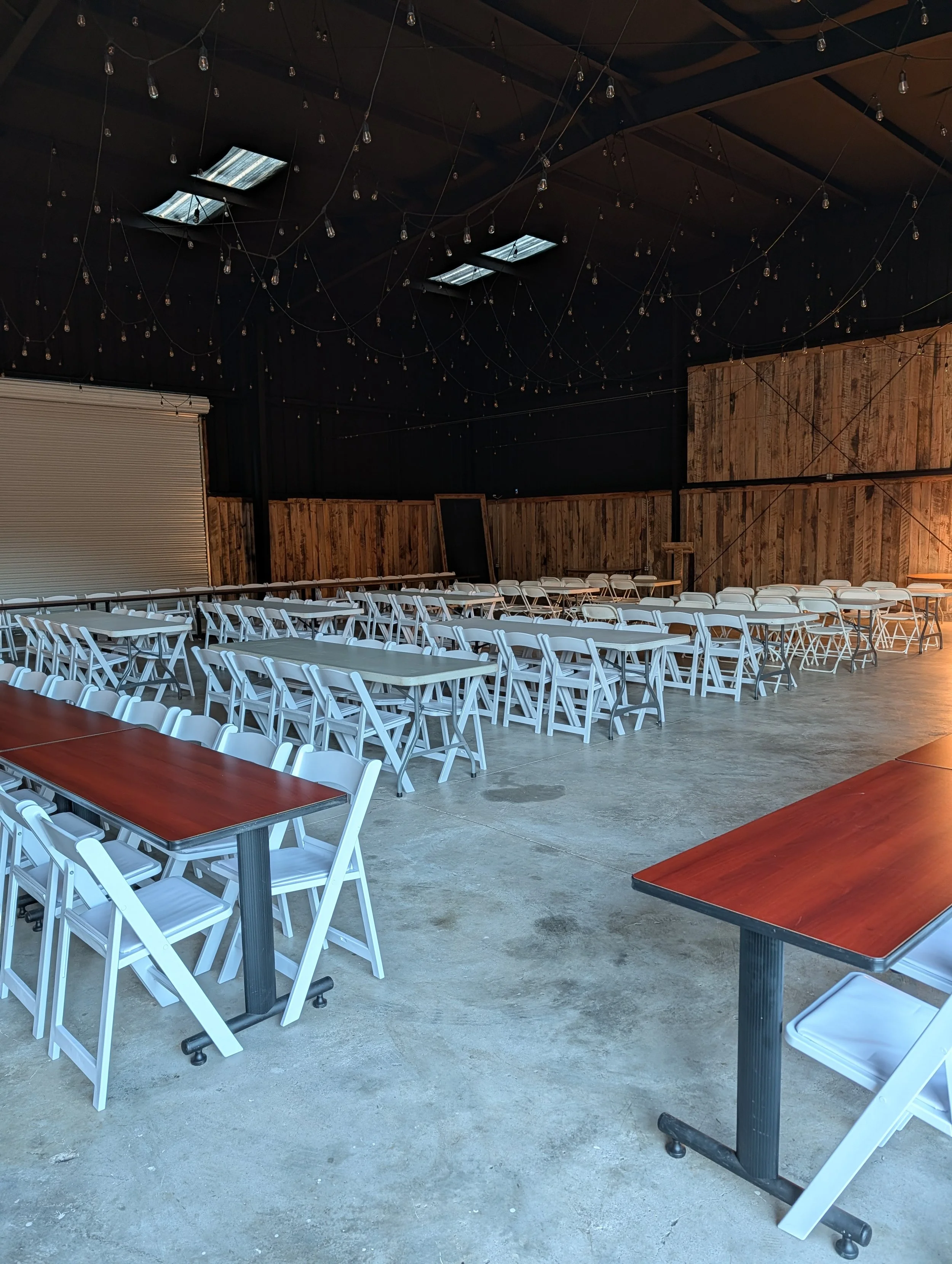 Empty event space with rows of white folding chairs and dark wood tables, hanging string lights, and a black ceiling with skylights.