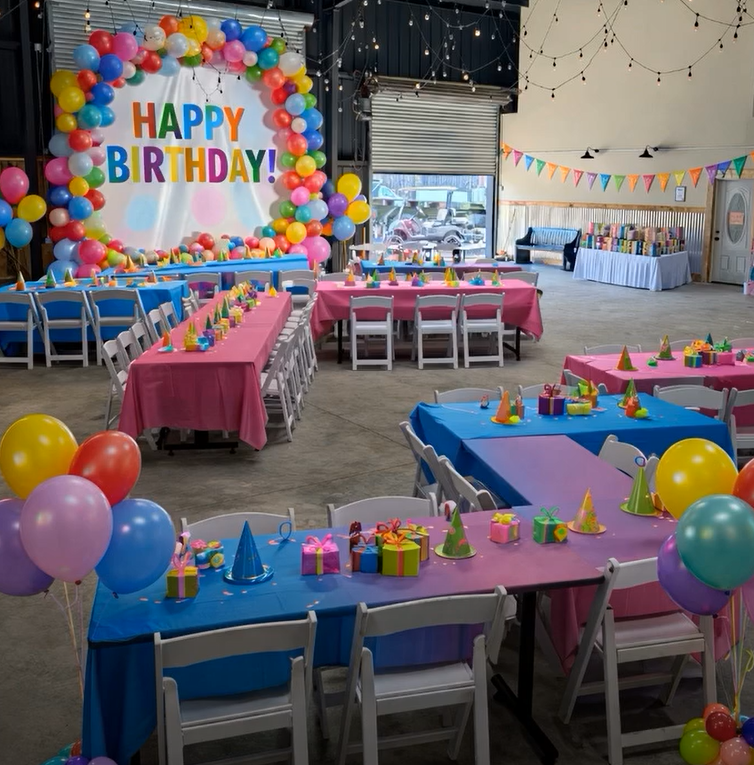 Indoor birthday party setup with colorful balloons, tables with blue and pink tablecloths, party hats, and gift boxes; a large balloon arch with a 'HAPPY BIRTHDAY!' banner as decor.