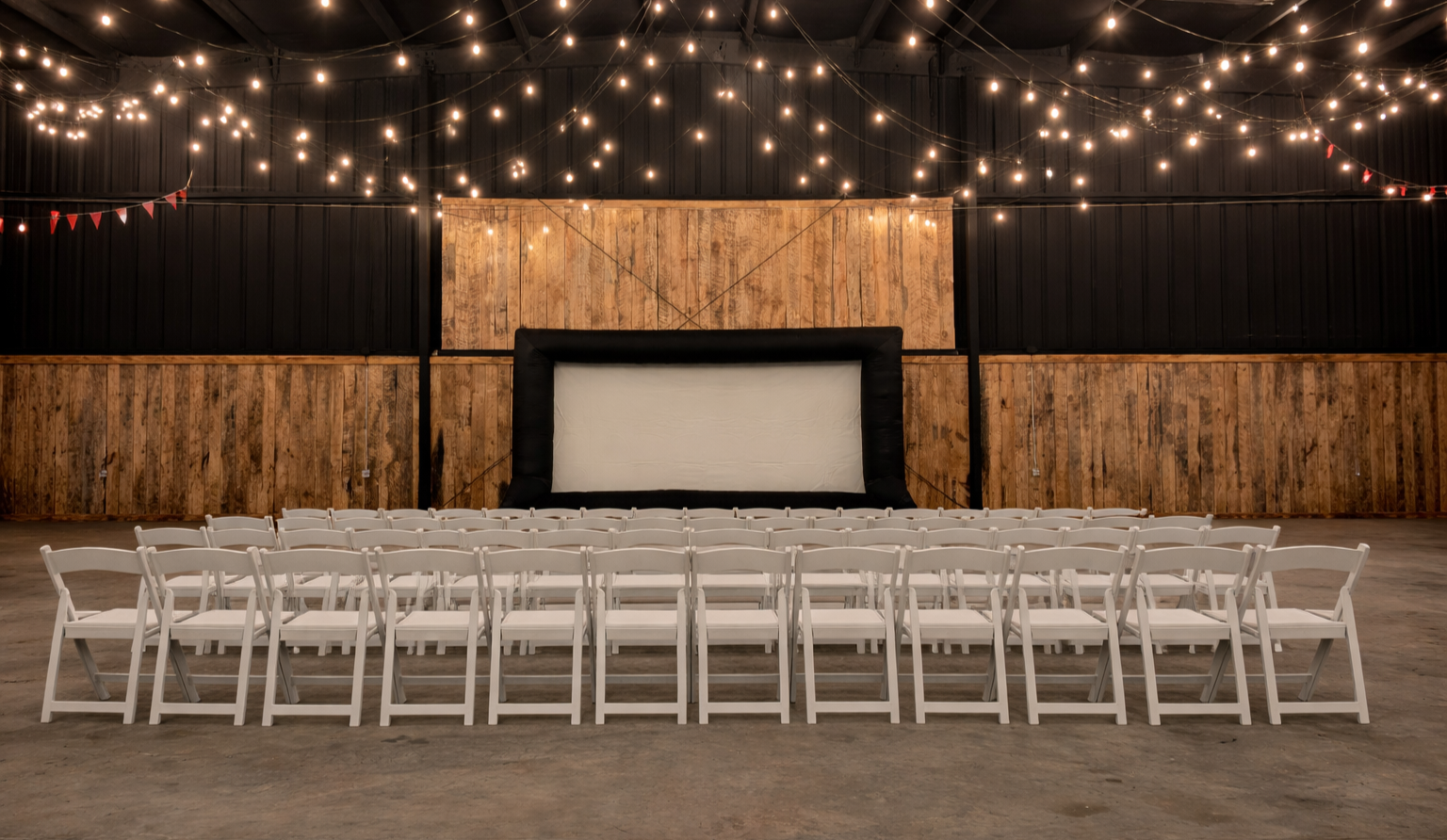 An empty indoor event space with white folding chairs arranged facing a large inflatable movie screen, decorated with hanging string lights and small red banners, with wooden walls and concrete floors.