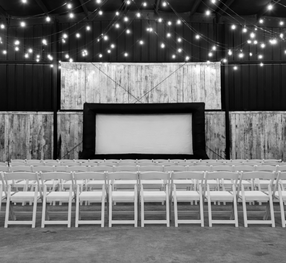 Empty indoor movie theater with rows of white chairs, a large screen, wooden walls, and string lights hanging from the ceiling.