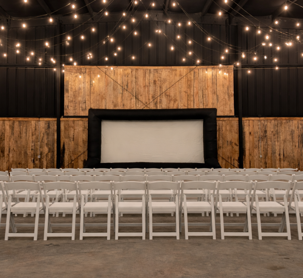 Empty chairs facing a large screen in a rustic indoor space with string lights overhead.