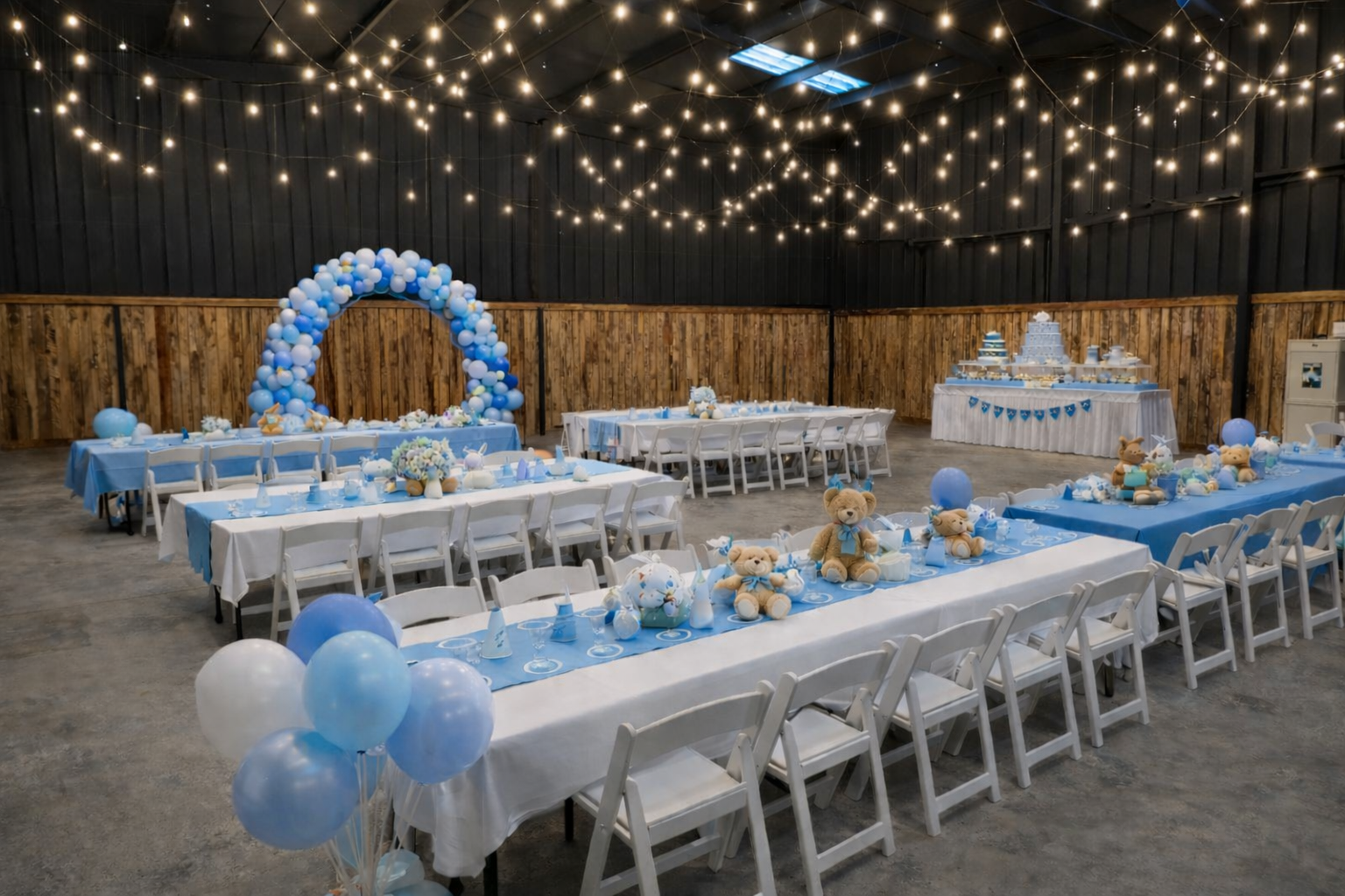 Indoor birthday celebration decorated in blue and white with teddy bears, balloons, a cake, and a balloon arch, featuring string lights hanging from the ceiling.