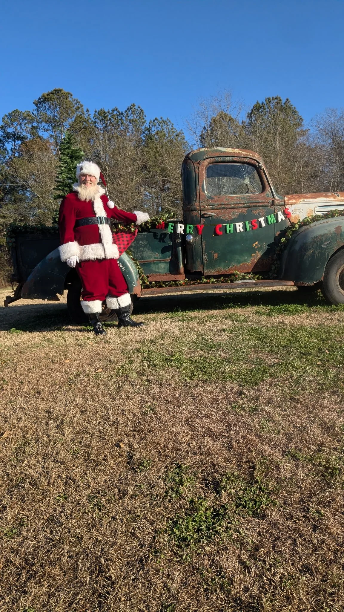 A person dressed as Santa Claus standing next to a rusty, vintage green truck decorated with a Christmas banner and greenery, with a clear blue sky and trees in the background.