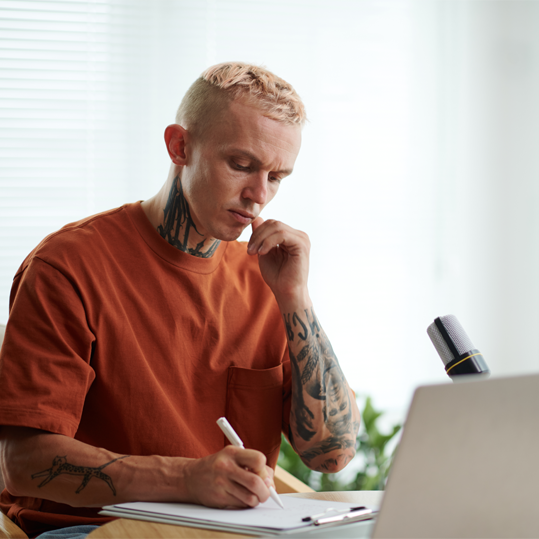Man with tattoos in a rust-colored t-shirt sitting at a desk, writing in a notebook, with a laptop and microphone nearby.