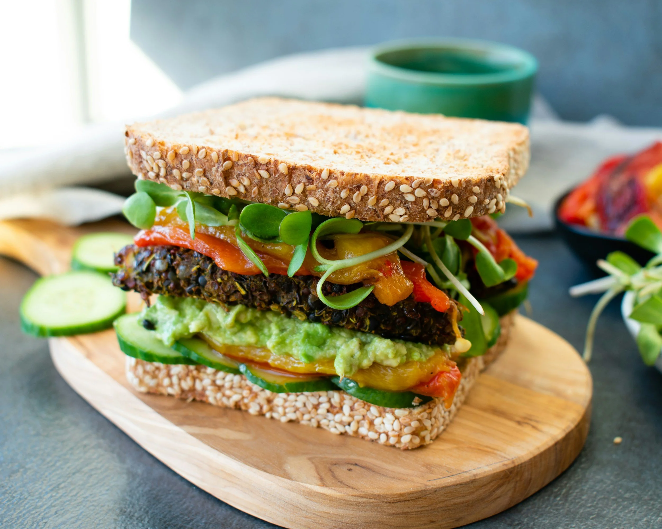 Close-up of a sandwich with sesame seed bun, meat patty, avocado, pickles, roasted peppers, sprouts, tomato, and lettuce on a round wooden board.