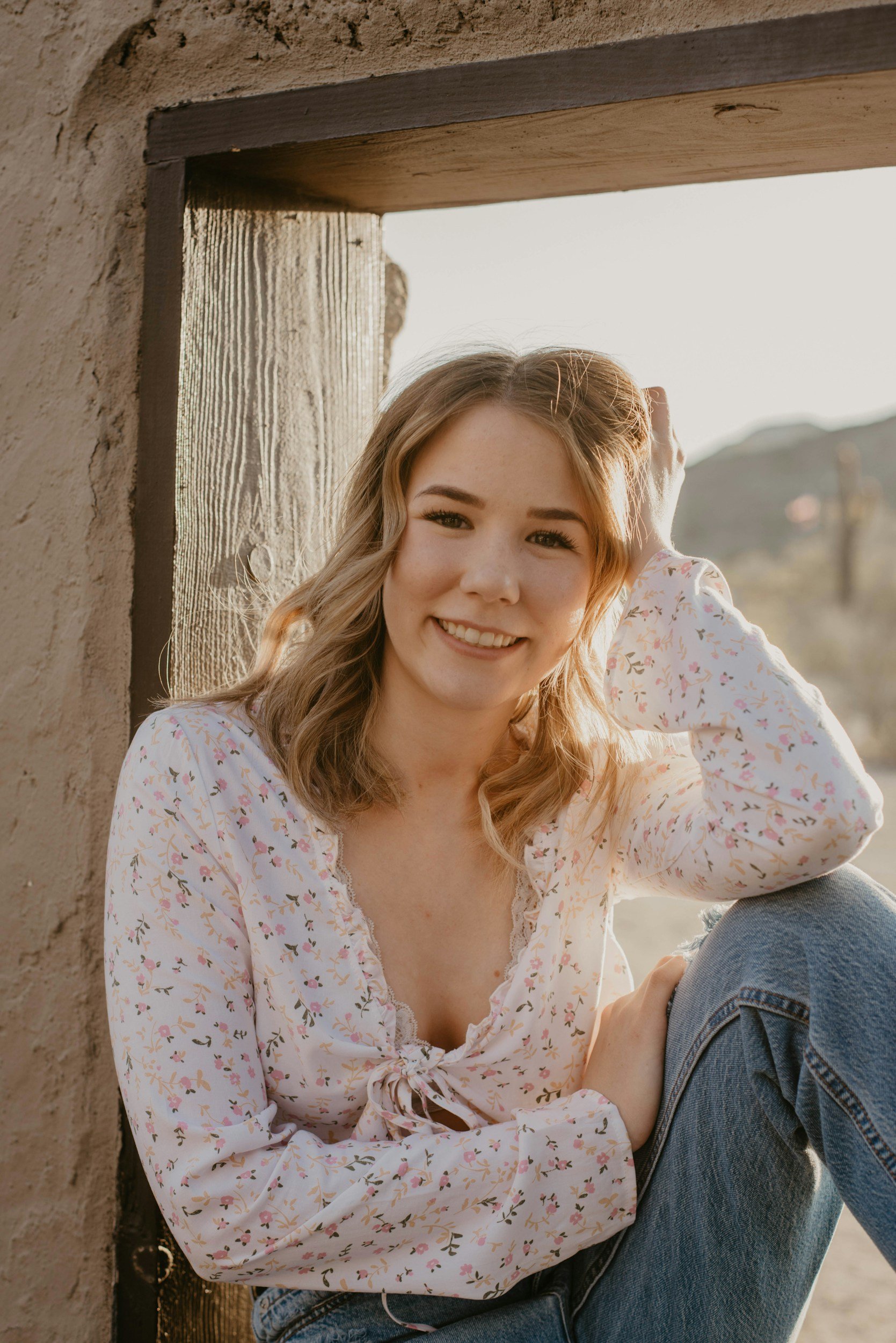 A young woman with light brown, wavy hair and fair skin smiling at the camera, sitting outdoors near a rustic wooden and stucco wall, wearing a white floral blouse and jeans.