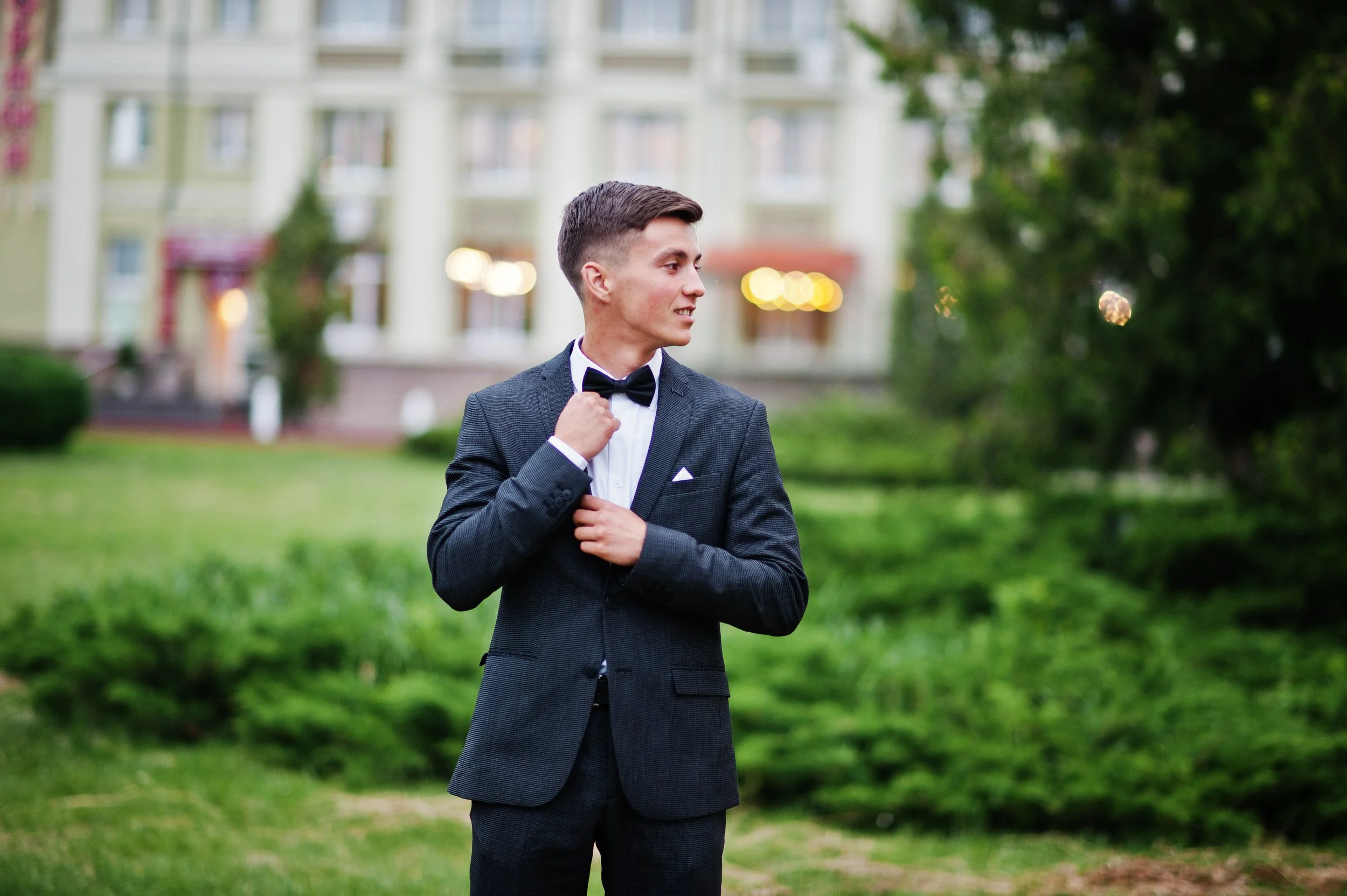 A young man in a black tuxedo with a bowtie stands outdoors on green grass, adjusting his bowtie and looking to his left, with a blurred building and trees in the background.