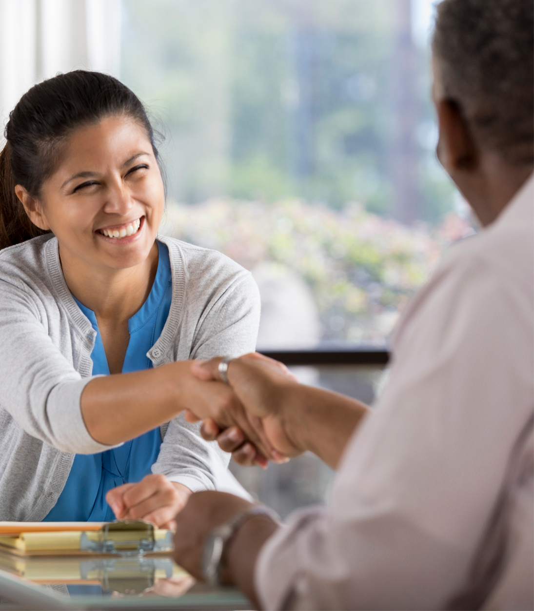 A woman smiling and shaking hands with a man in an indoor setting with a window behind them.