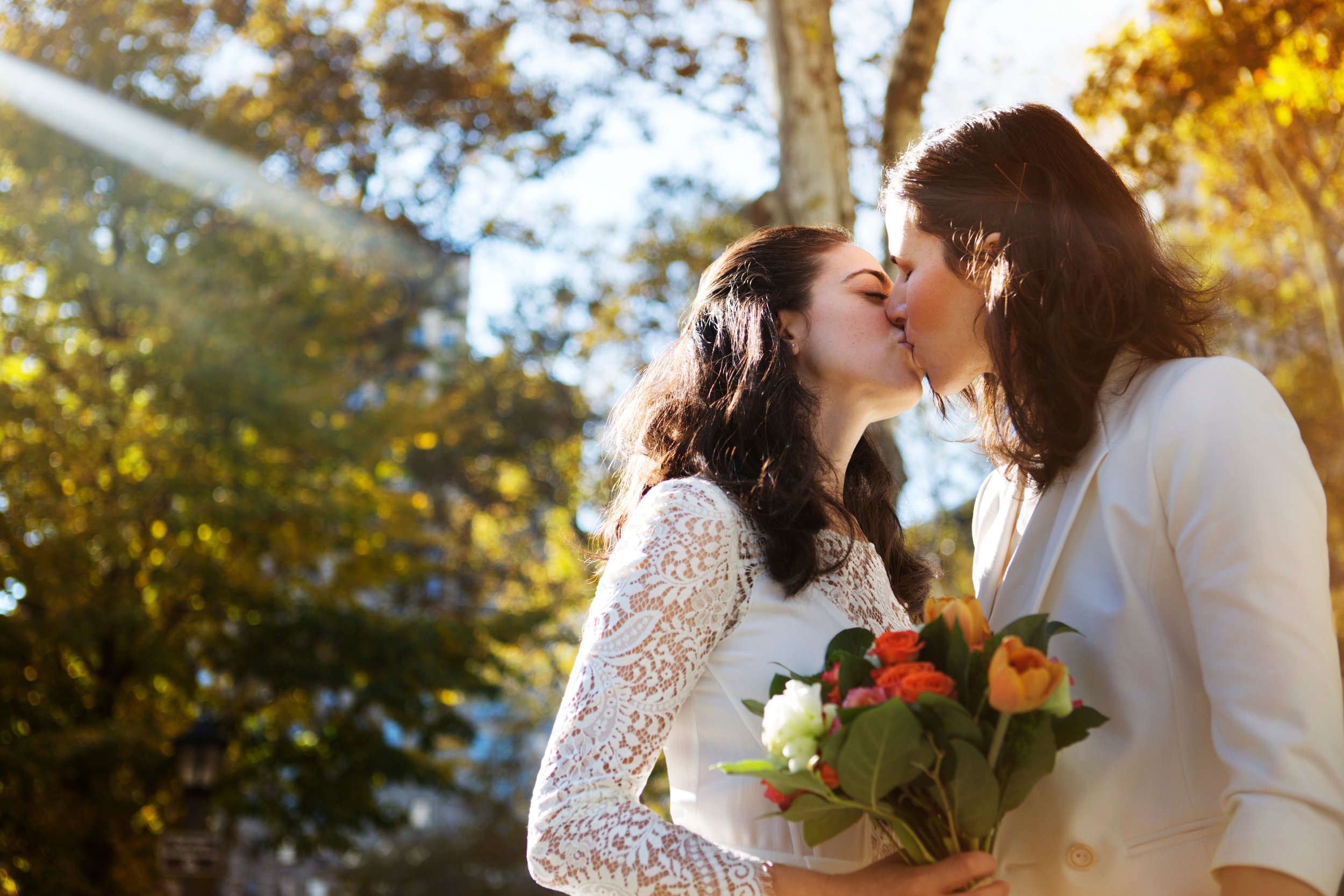 Two women sharing a kiss during their outdoor wedding on a sunny day, with one holding a bouquet of flowers, trees with autumn leaves in the background.