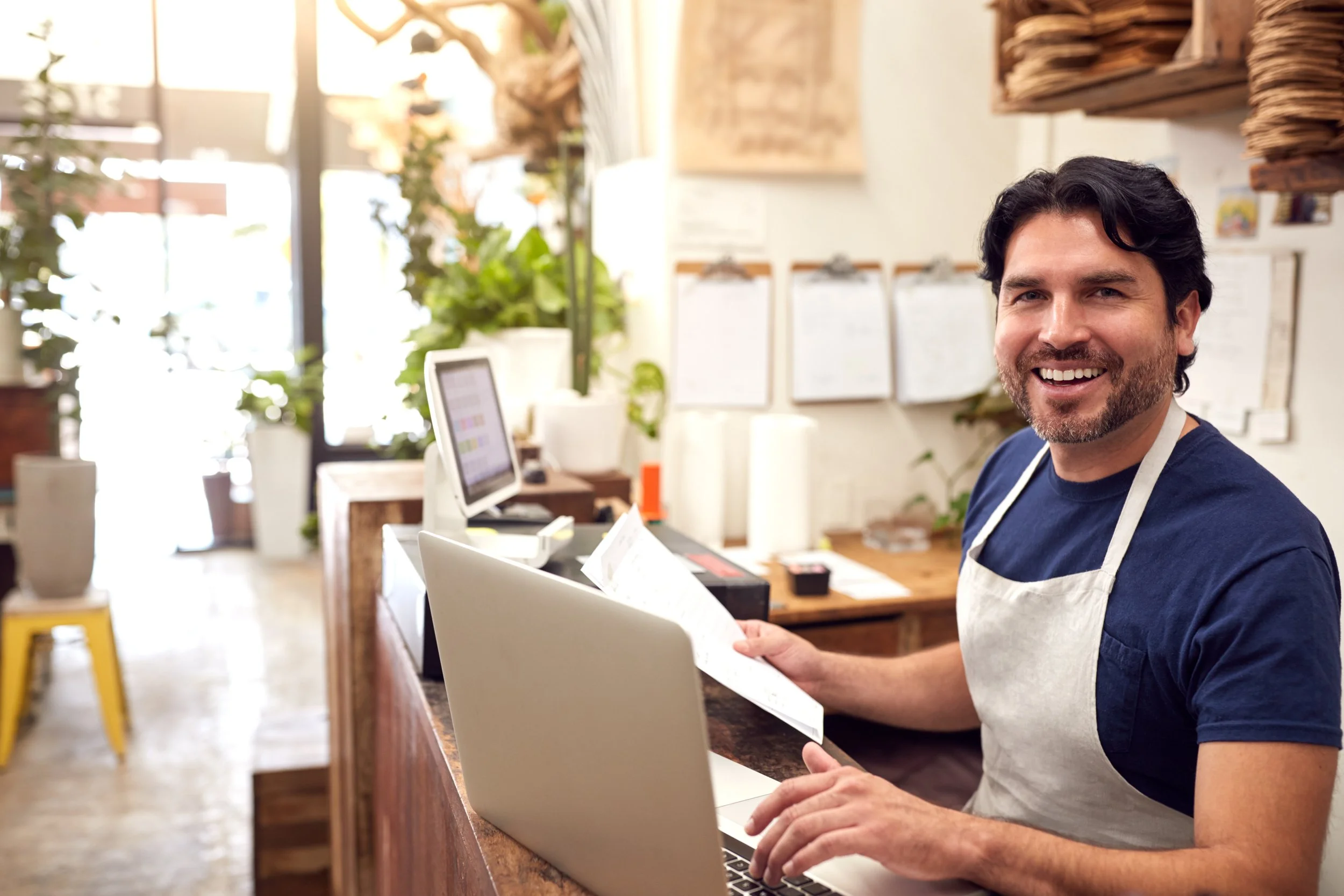 Image shows a smiling small business owner holding an invoice while using a laptop to look at their business website