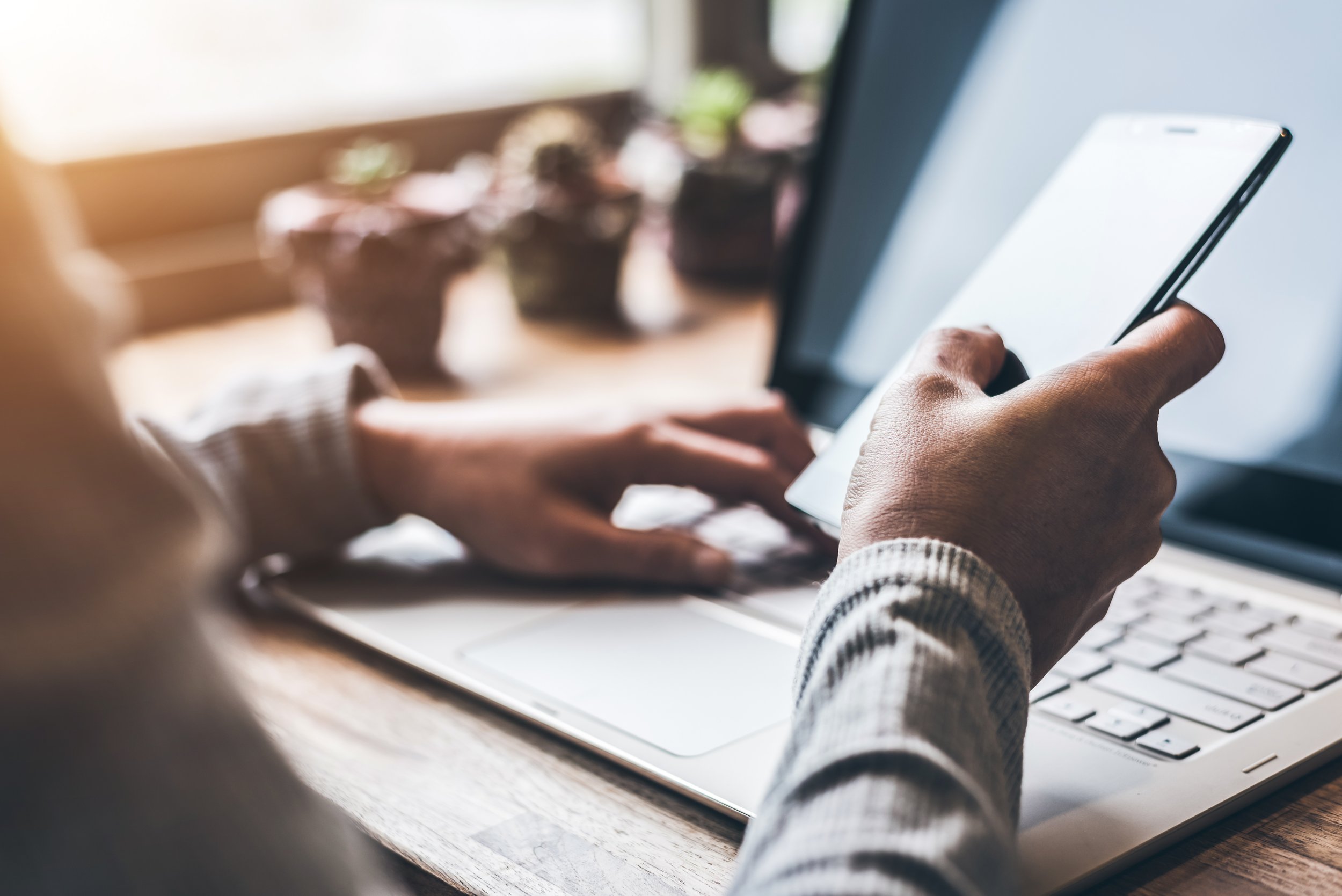 Image shows person holding a cell phone while looking at a laptop.